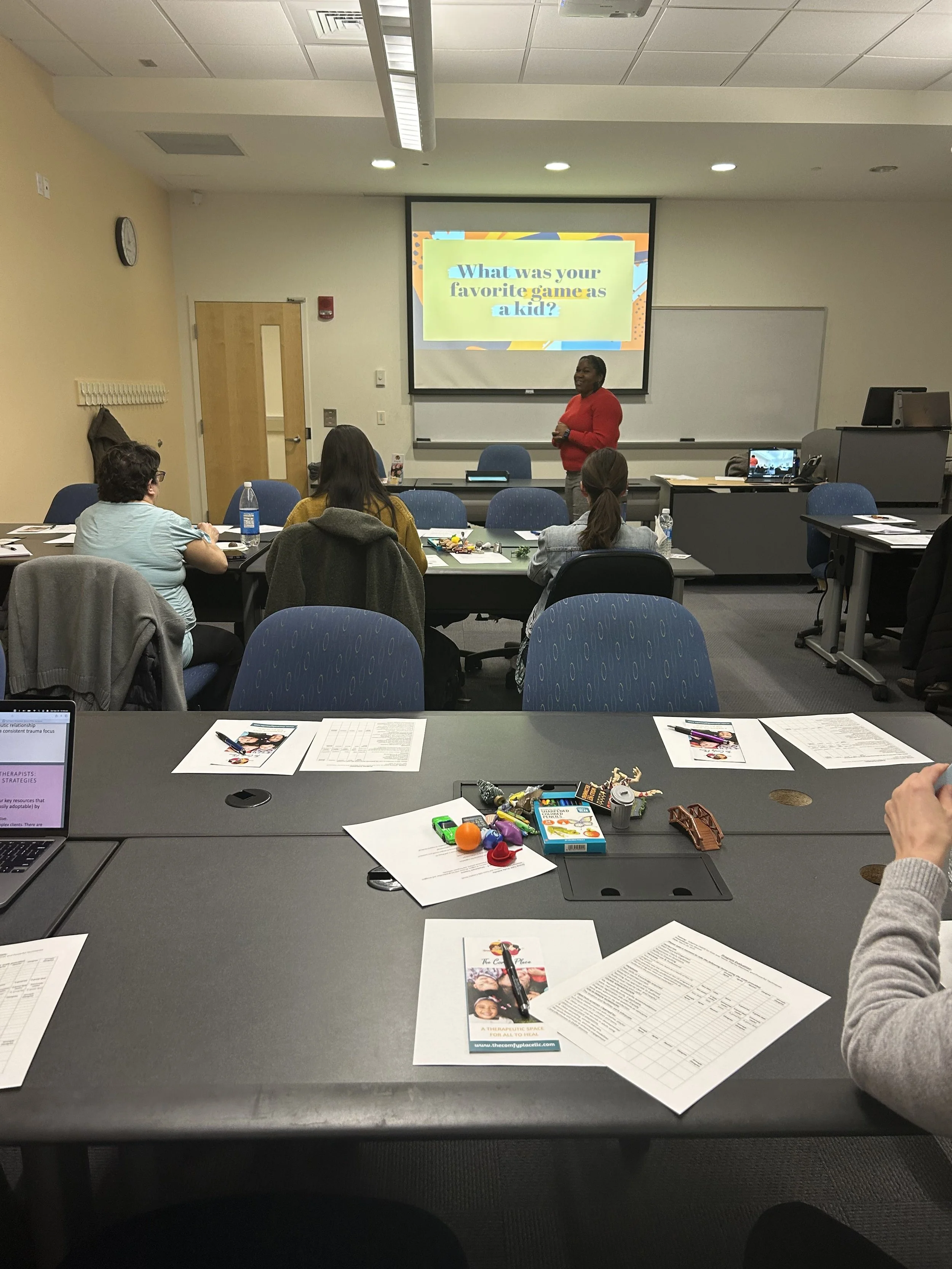 A classroom or seminar room with a presenter standing in front of a screen, which displays a slide asking "What was your favorite game as a kid?". Several attendees sit facing the presenter at tables, with papers, a laptop, and various small objects 