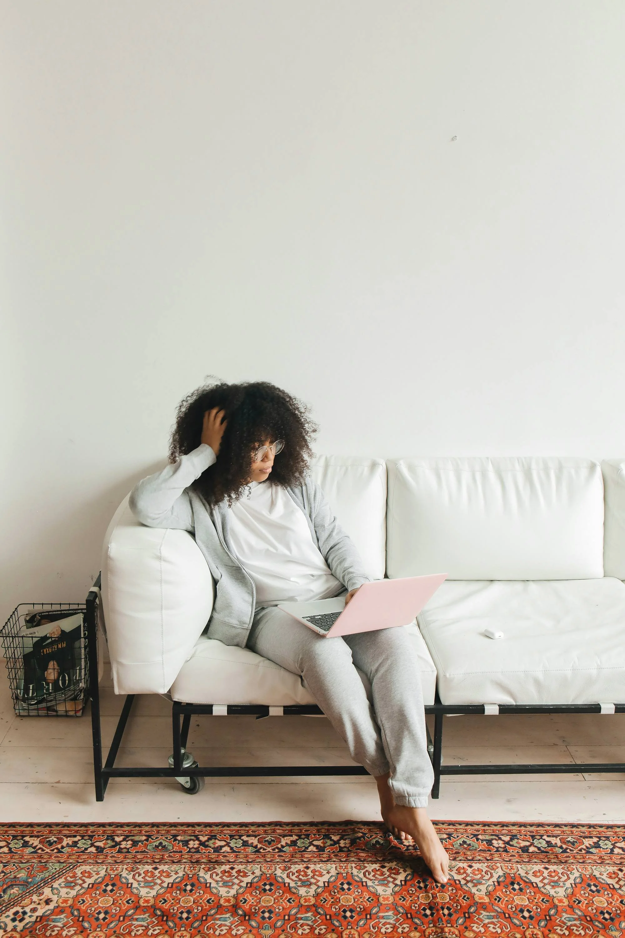 A woman with curly hair and glasses sitting on a white couch, using a pink laptop, with a magazine and a magazine rack nearby on a hardwood floor. She is wearing a gray hoodie and sweatpants and appears to be in a relaxed setting.