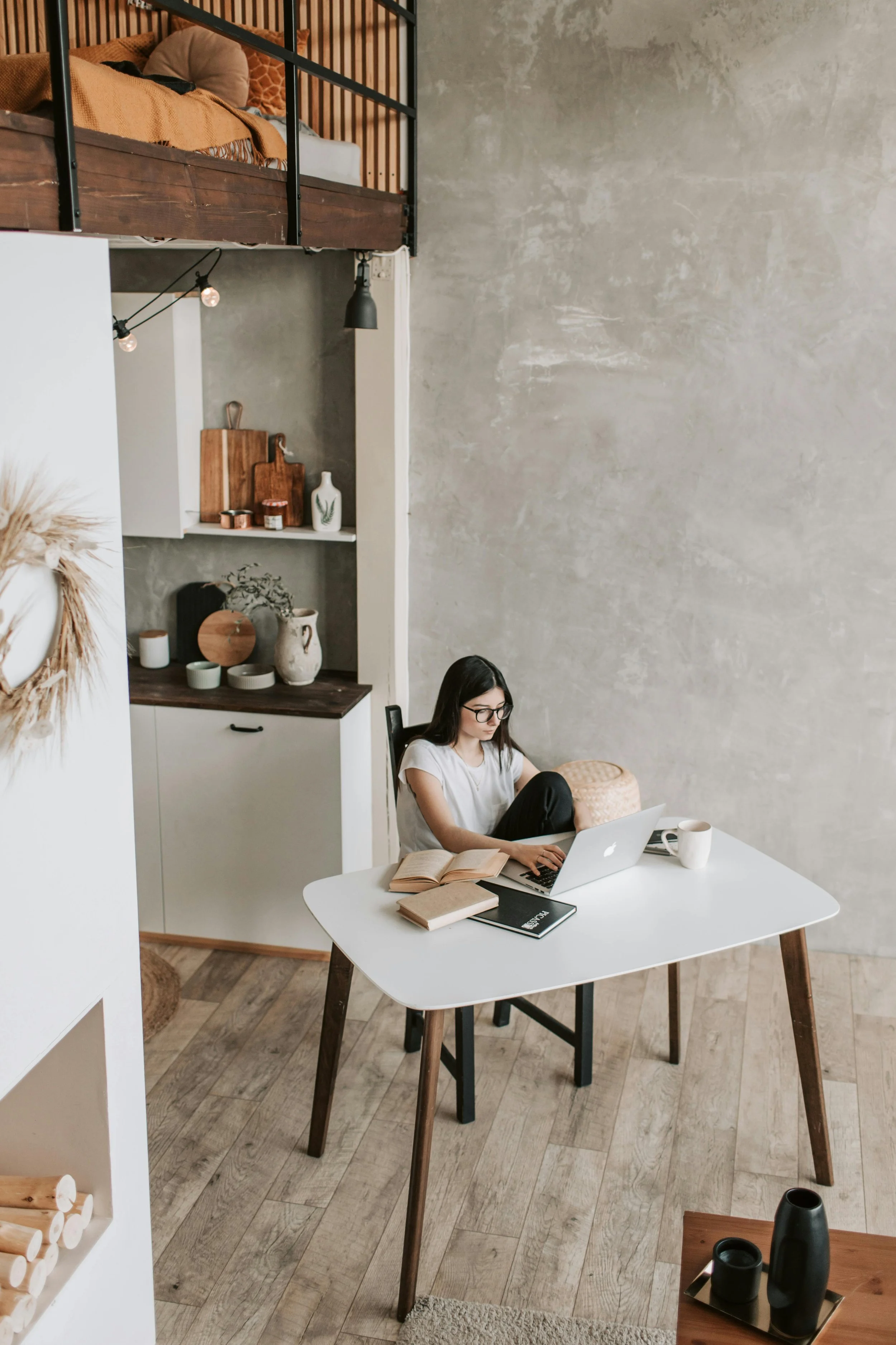A woman with glasses sitting at a white table using a laptop, with notebooks and a book in front of her, in a modern, minimalist home with wood and neutral tones.