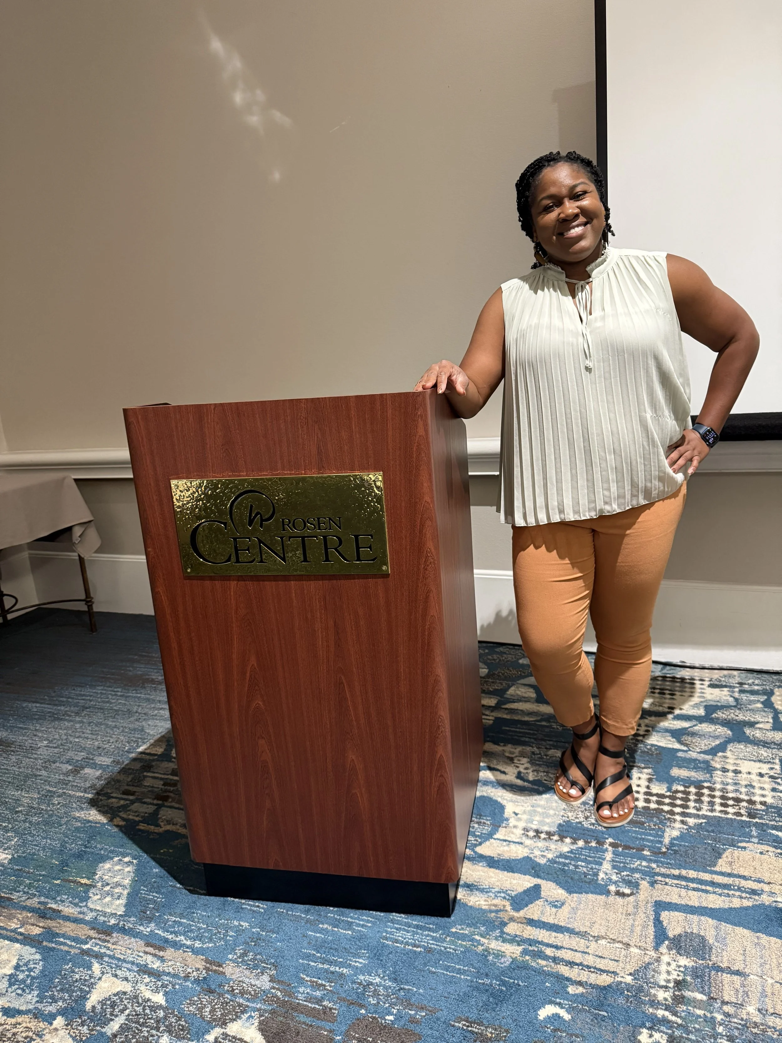 A woman, Lisa Ibekwe, in a sleeveless cream blouse and tan pants standing next to a wooden podium labeled 'Rosen Centre', smiling in a conference room with a patterned carpet and a blank wall.
