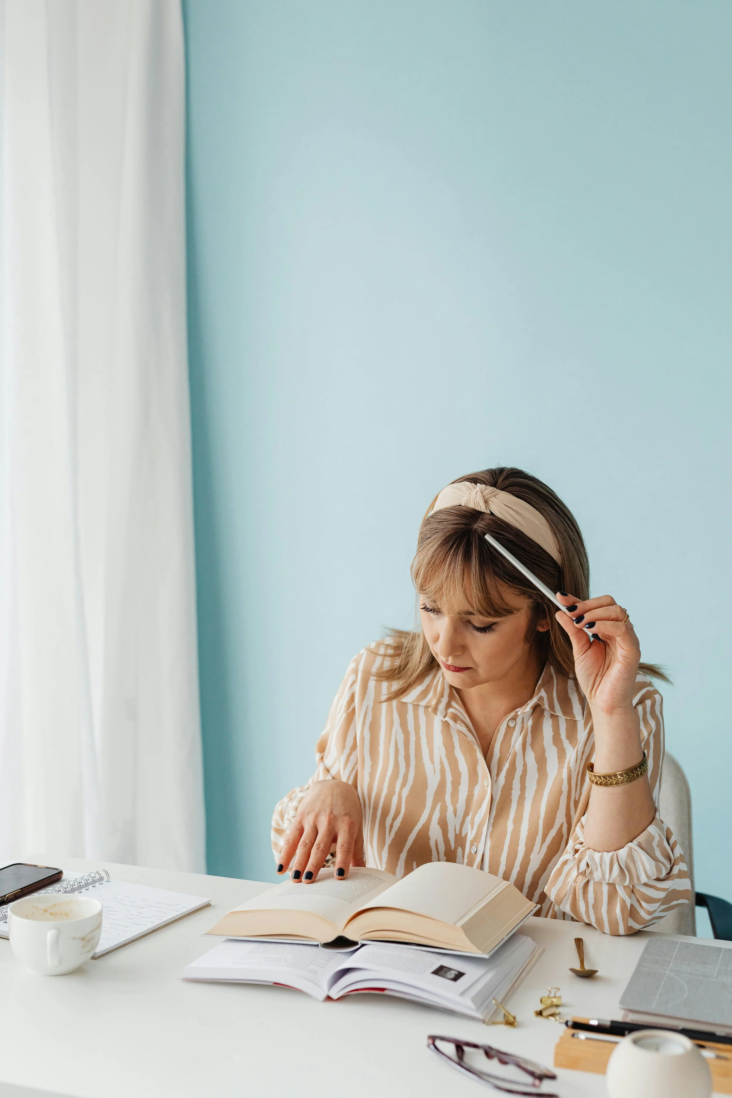 A woman with tan hair, wearing a beige and white striped shirt, a beige headband, and a gold bracelet, studying with open books and notebooks on a white desk in front of her, holding a pen to her forehead.