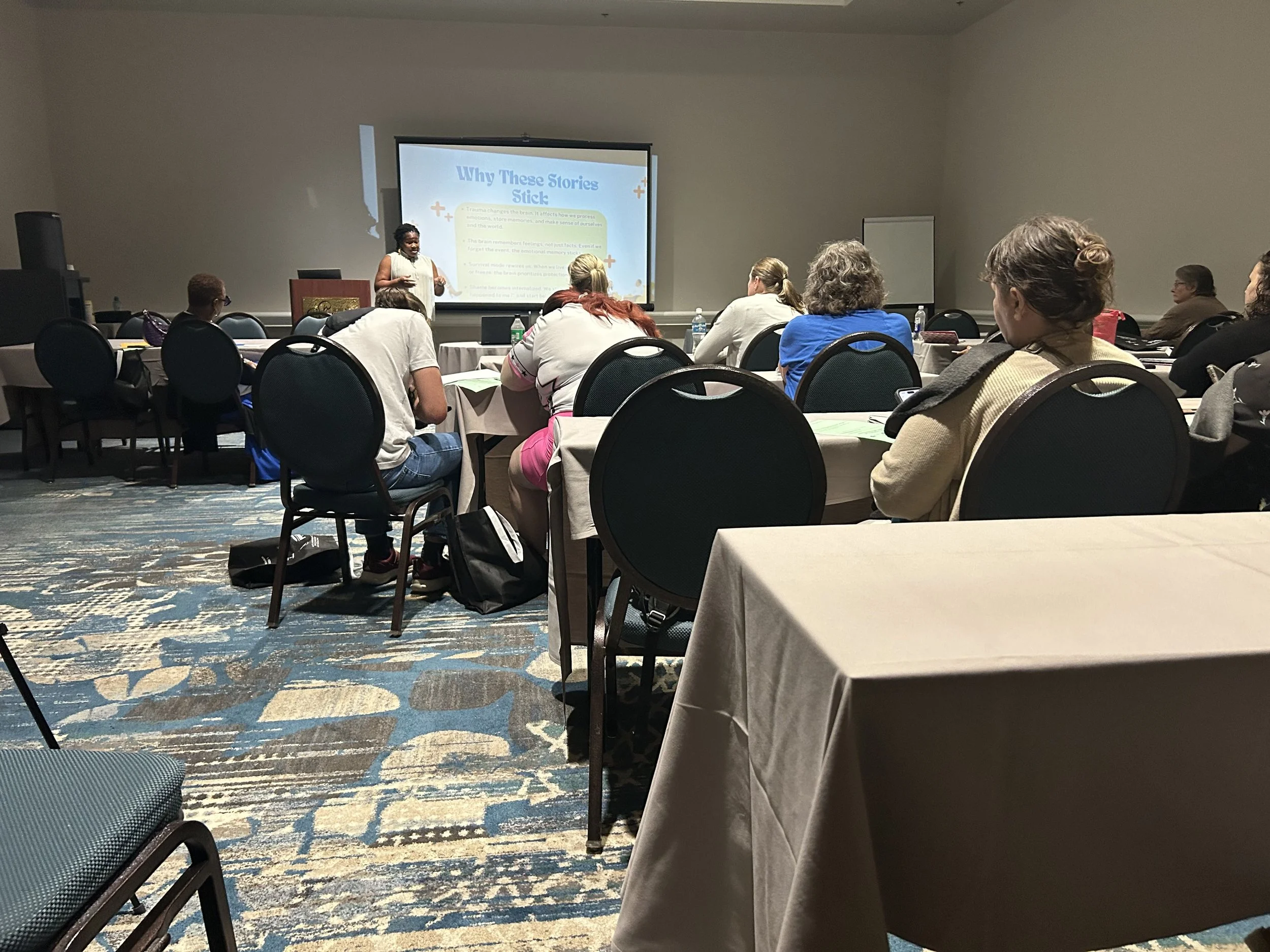A group of people seated in a conference room, watching a presentation on a screen. The presenter, Lisa Ibekwe, stands at the front near the screen with a slide displaying the title 'Why These Stories Stick'.