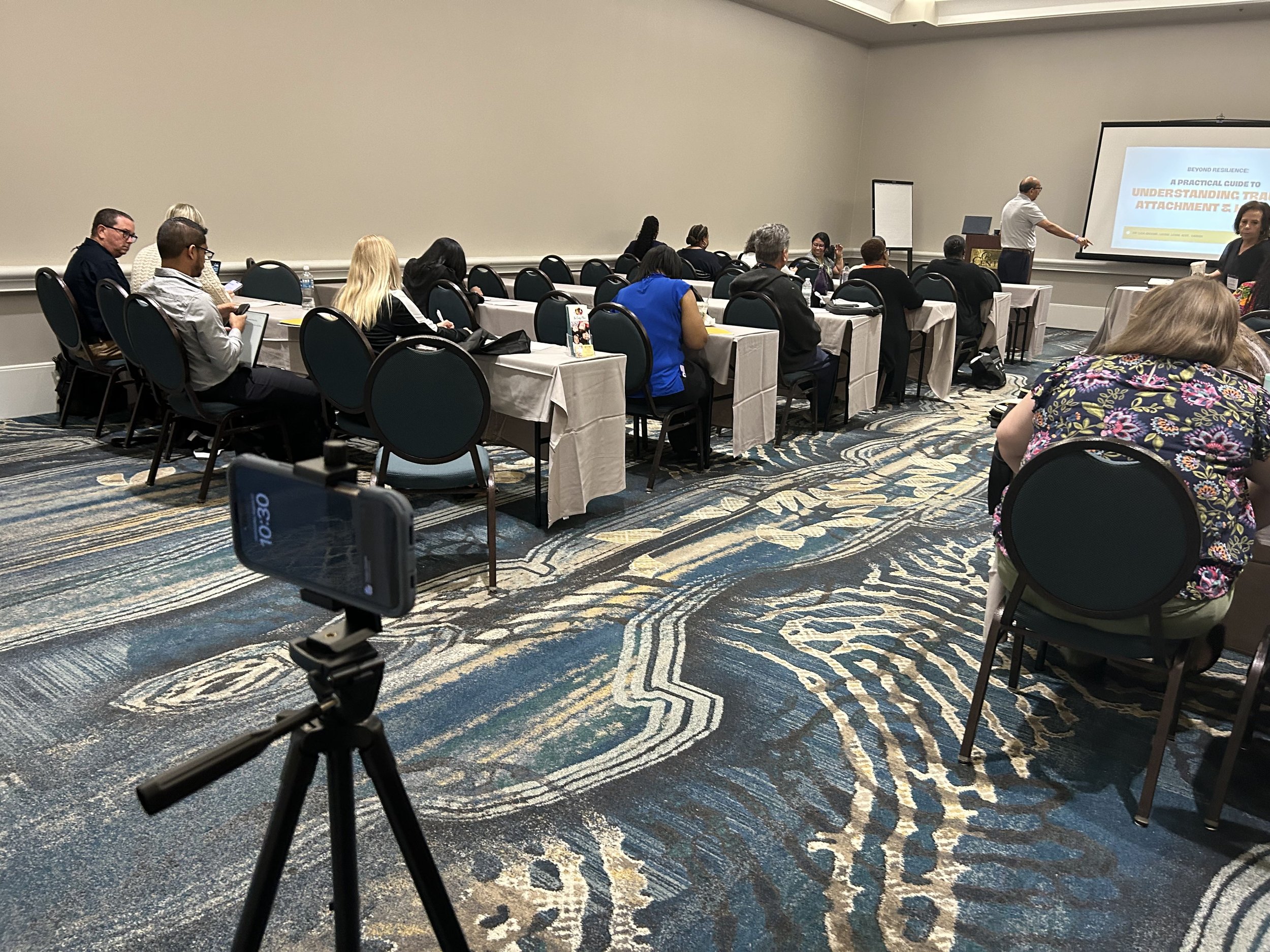 A conference room with people seated at tables listening to a presentation. A man is at the front pointing to a slide on a large screen, which reads 'A Practical Guide to Understanding Tra... Attachment 2'. A camera on a tripod is set up in the foreg