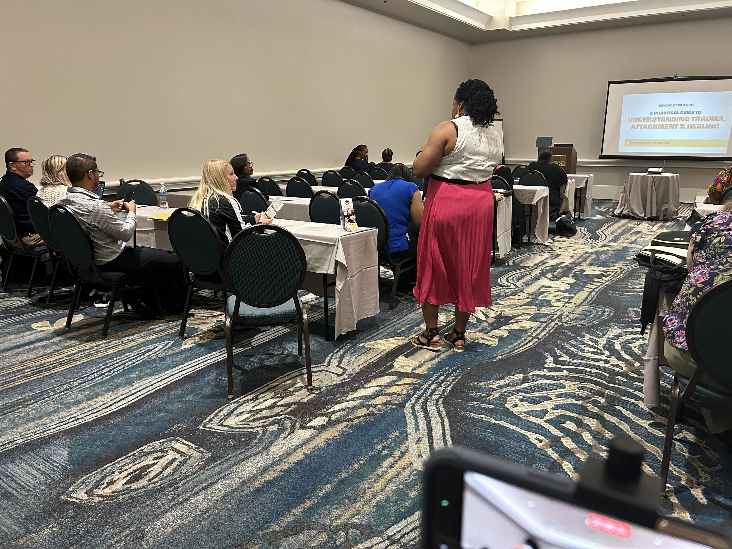 A woman, Lisa Ibekwe, in a white top and pink skirt stands in front of a seated audience in a conference room, facing a presentation on understanding trauma, attachment, and healing displayed on a large screen.