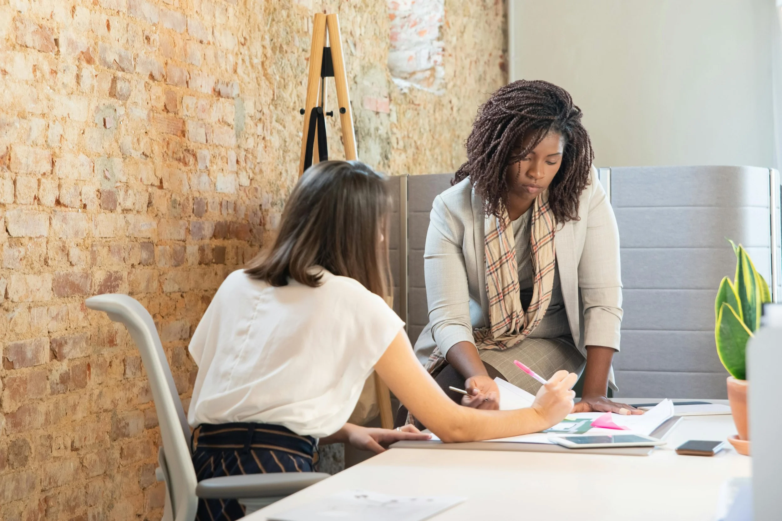 Two women having a discussion at a desk in an office with a brick wall background.