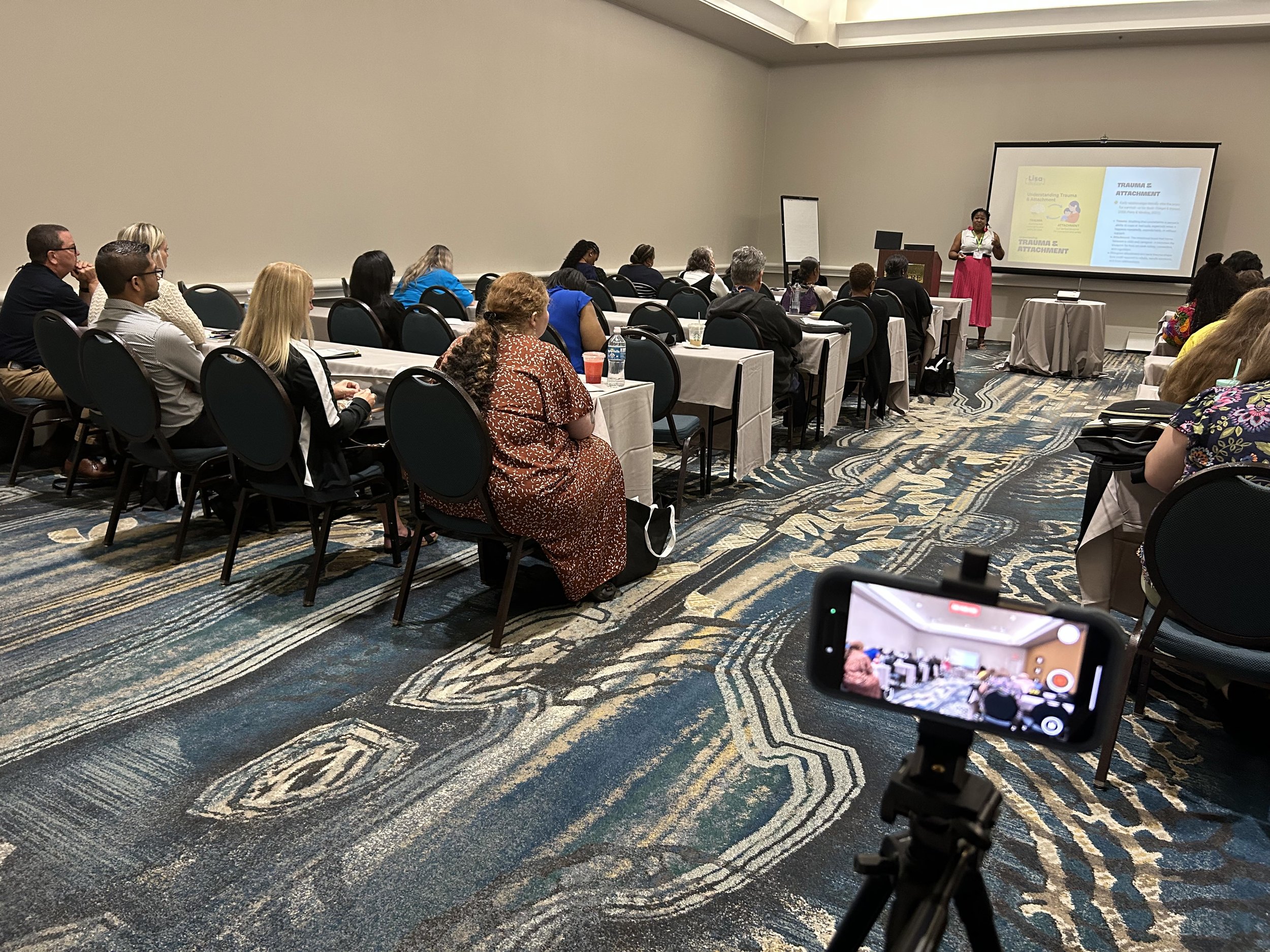 A woman, Lisa Ibekwe, giving a presentation at a conference with a projected slide titled 'Trauma & Attachment'. Attendees sitting at tables listening to the speaker, with a camera recording the event in the foreground.
