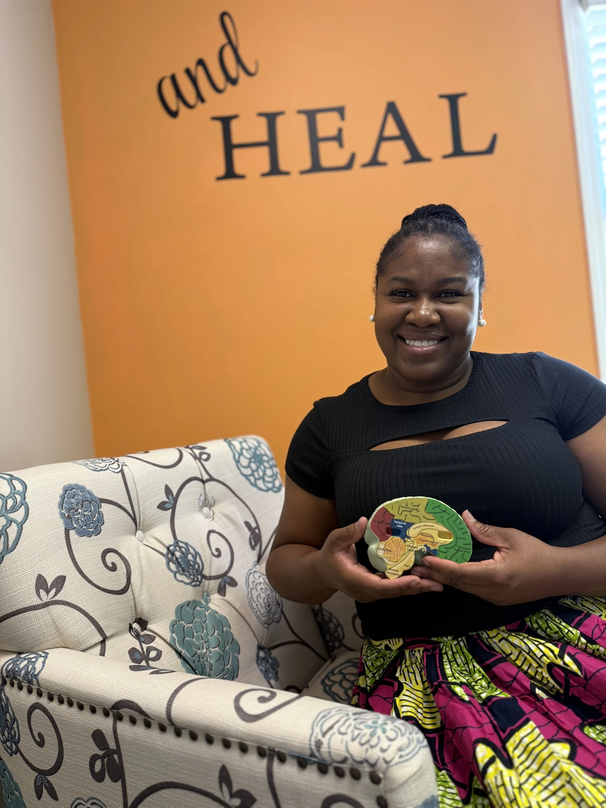 A smiling woman sitting on a floral upholstered couch holding a model of a human brain, with an orange wall behind her that has the words 'and HEAL' written on it.