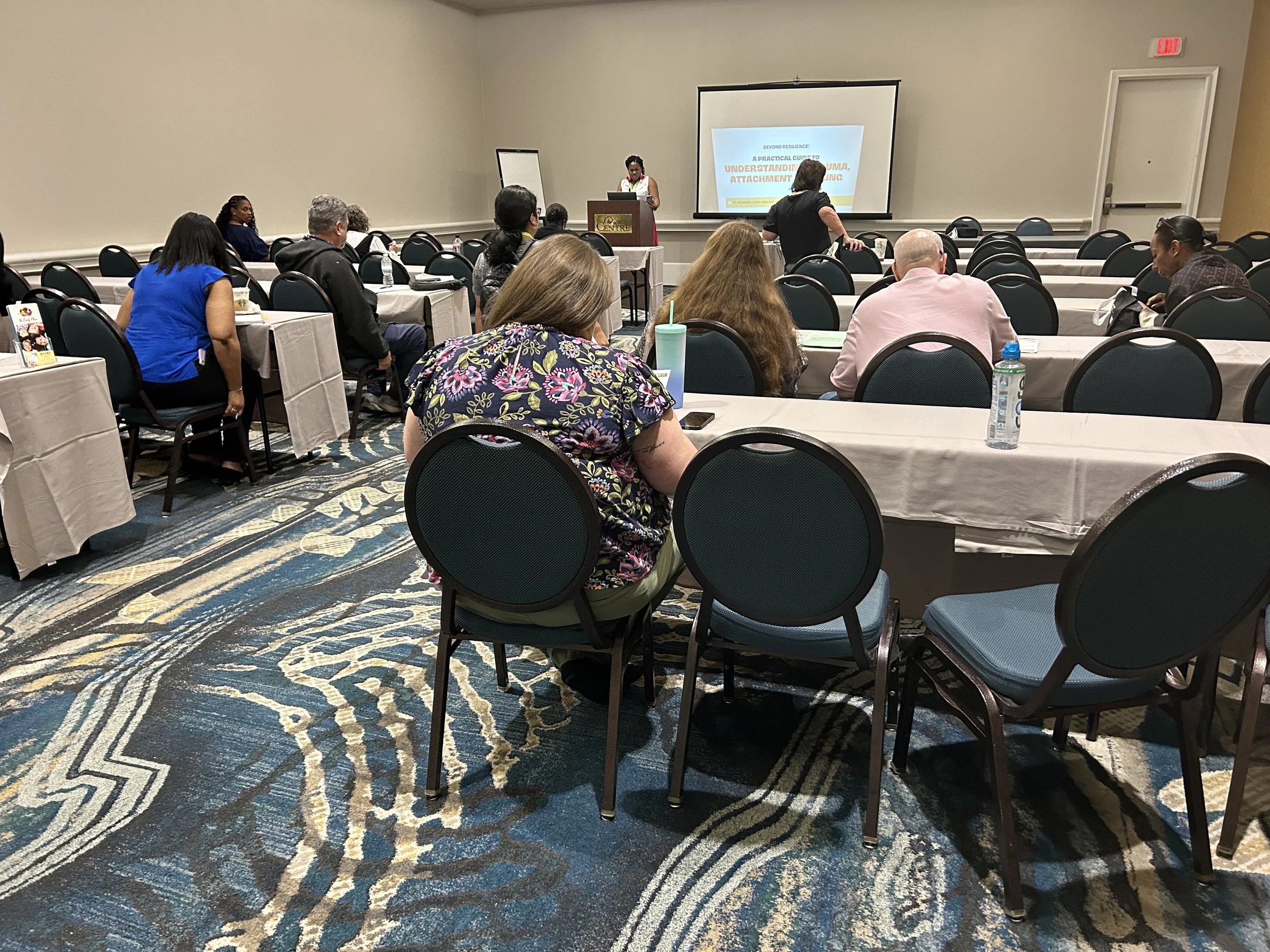 A seminar or conference room with attendees sitting at tables, facing a speaker at the front. There is a woman standing near the podium, presenting a slide on the screen that reads 'Understanding Attachment'.
