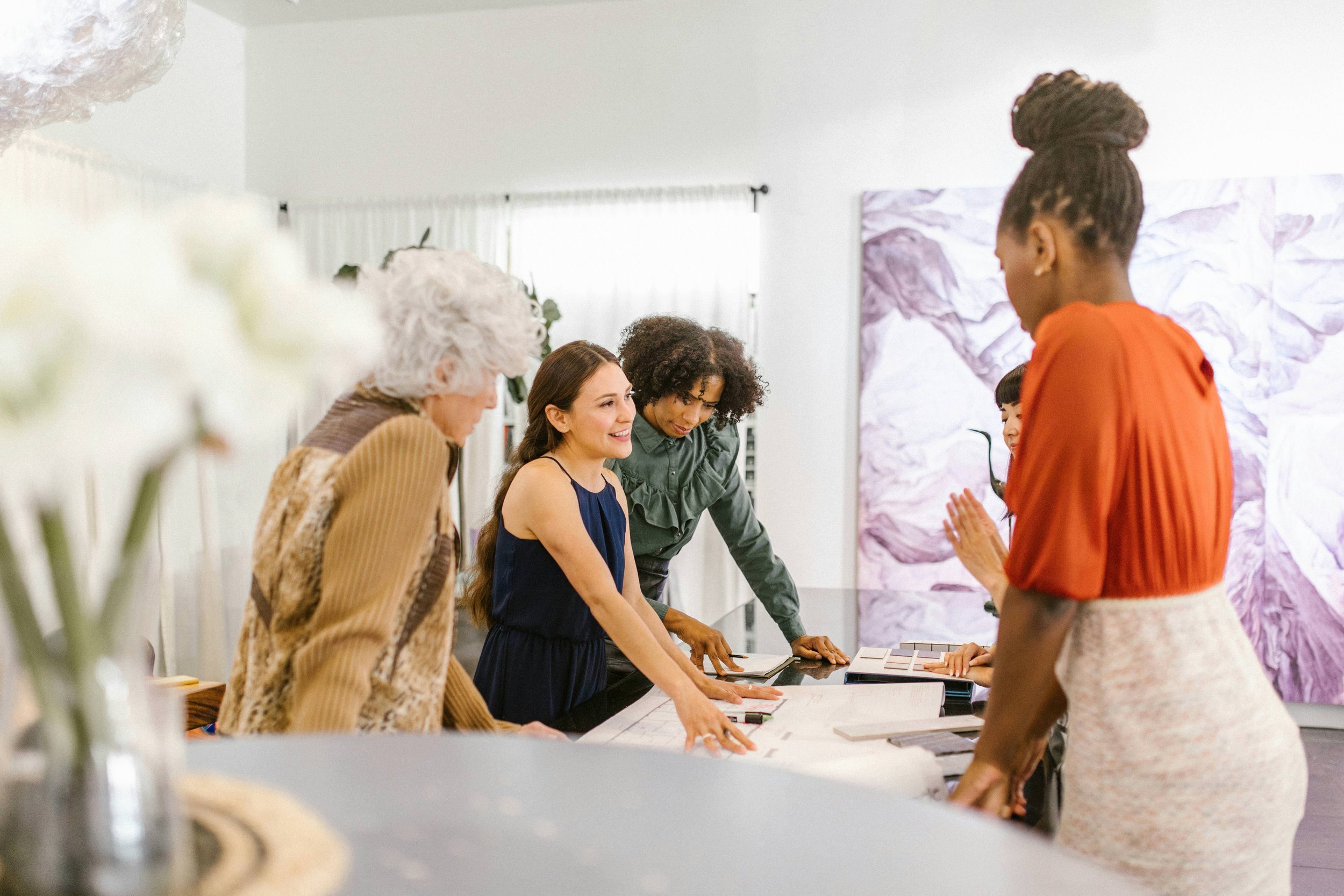 A diverse group of women gathered around a table, engaged in conversation and discussing fabric or design samples in a bright, modern room.
