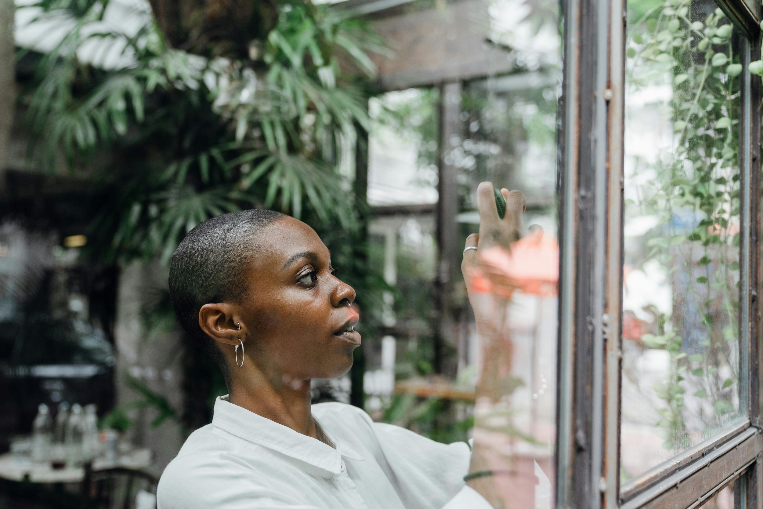 Woman with short hair and earrings looking through a glass window in a green, indoor setting.
