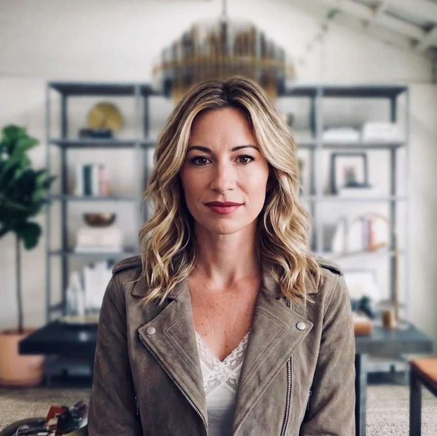 A woman with wavy blonde hair posing in a modern, well-lit office or living room with bookshelves behind her.
