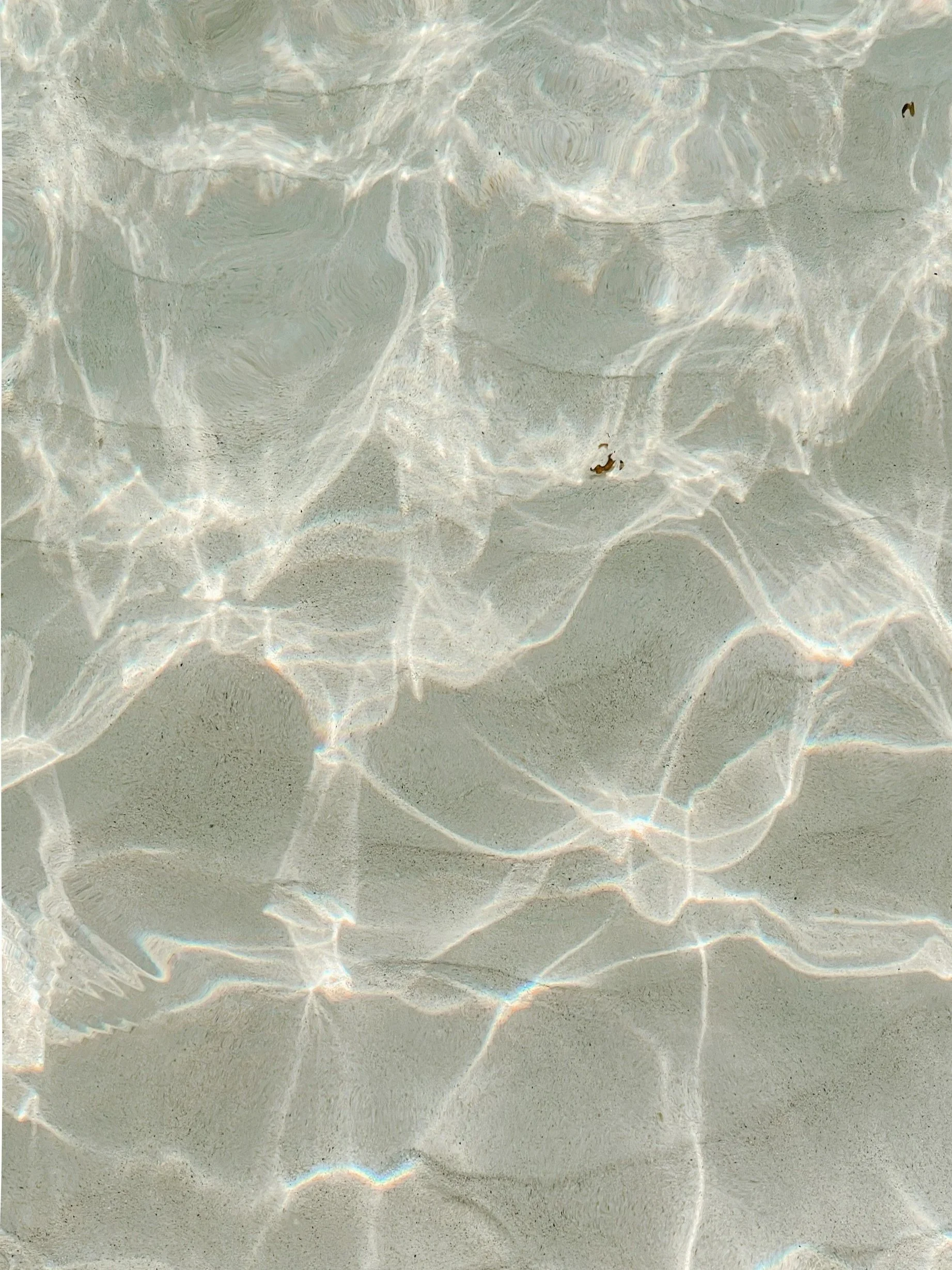 Shallow clear water with sunlight reflections and shadows on a sandy bottom.