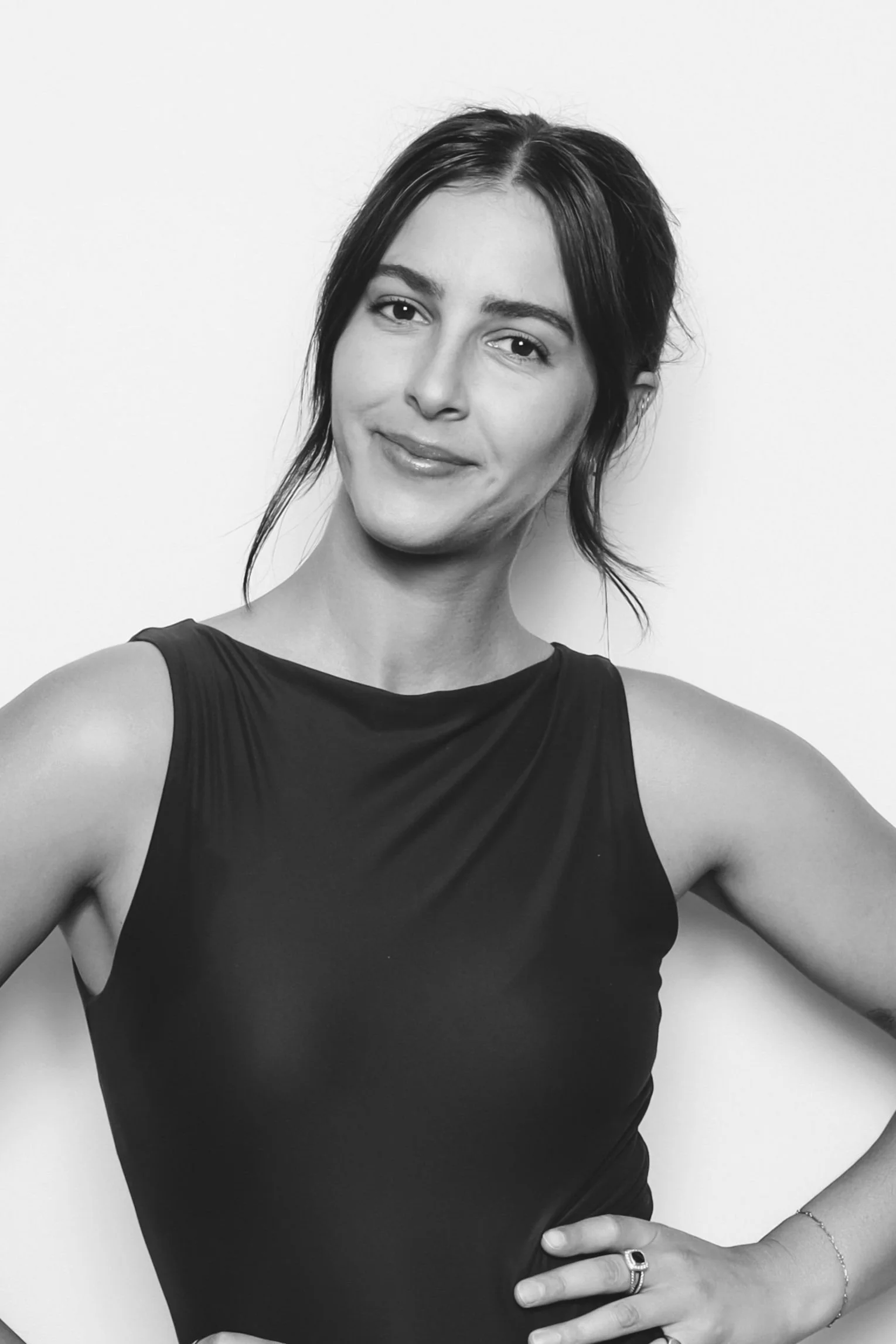 Black and white portrait of a woman with a slight smile, wearing a sleeveless top, with her right hand on her hip, standing against a plain background.