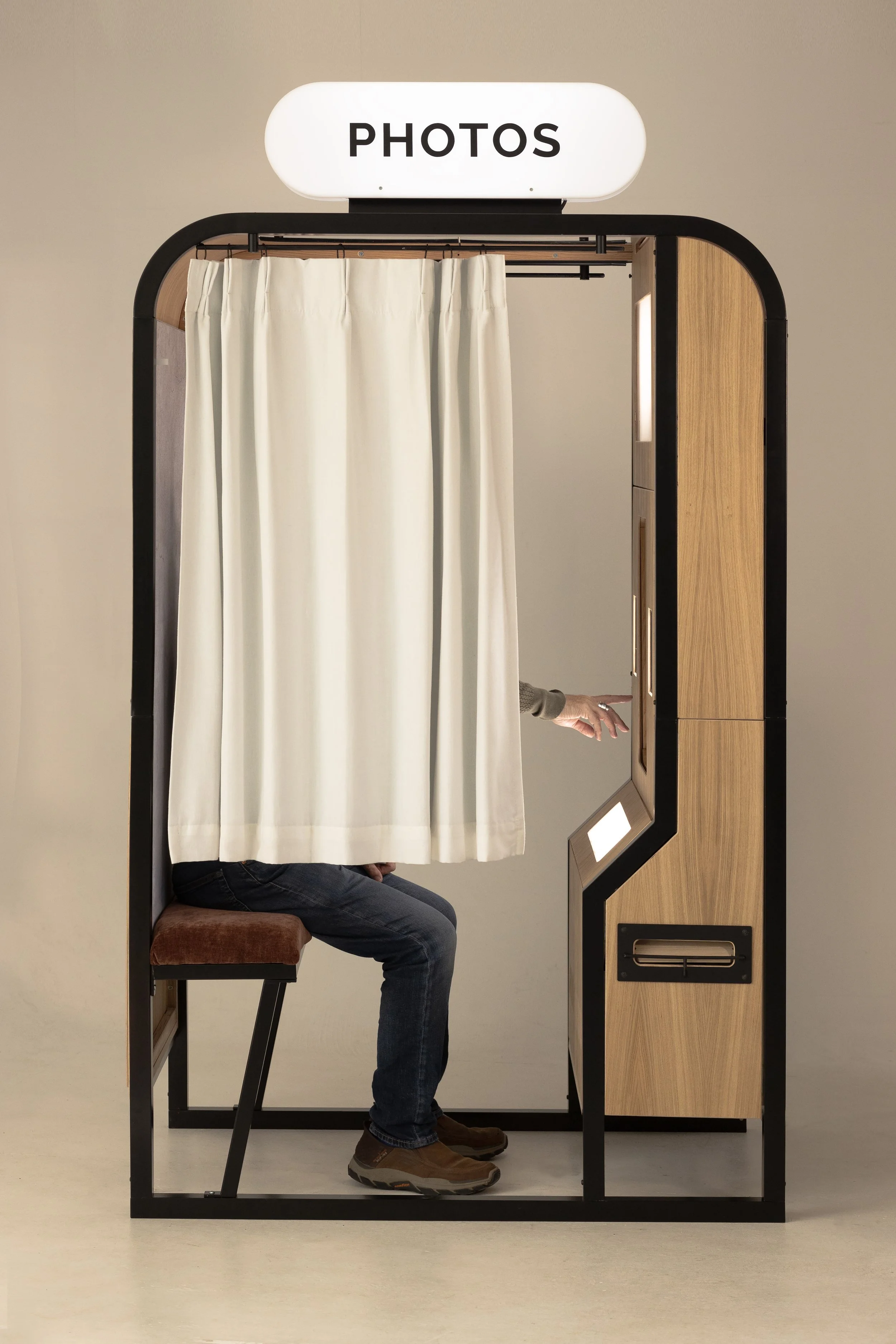 A person sitting on a brown cushioned stool inside a photo booth with a white curtain, wooden exterior, and a labeled "PHOTOS" sign on top, with someone else reaching through the booth's inside to operate the camera or controls.