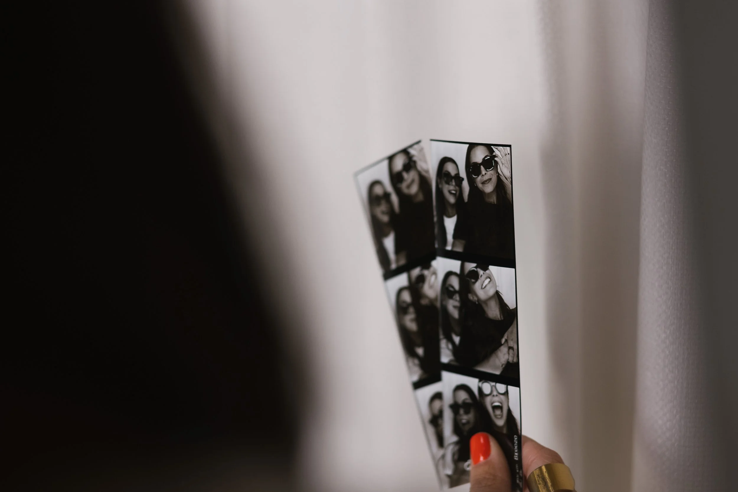 Close-up of a hand holding a strip of six black-and-white photo booth pictures of women wearing sunglasses and making playful faces against a white wall.