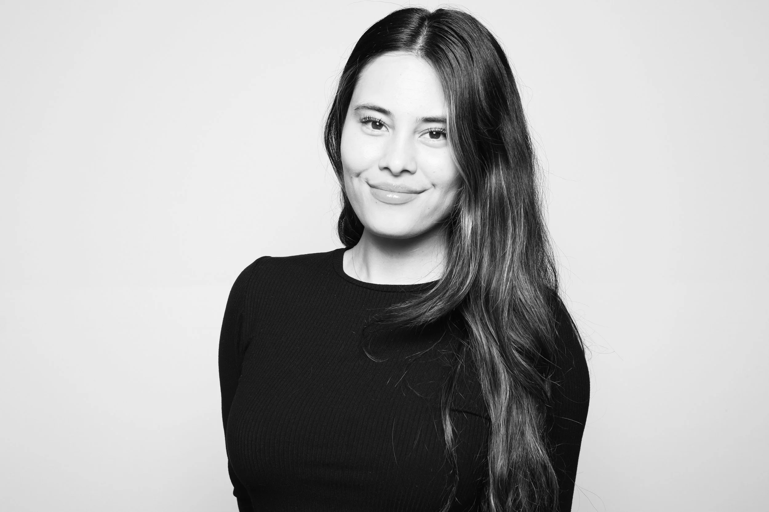 Black and white portrait of a young woman with long, wavy hair wearing a black top, smiling slightly while looking at the camera.
