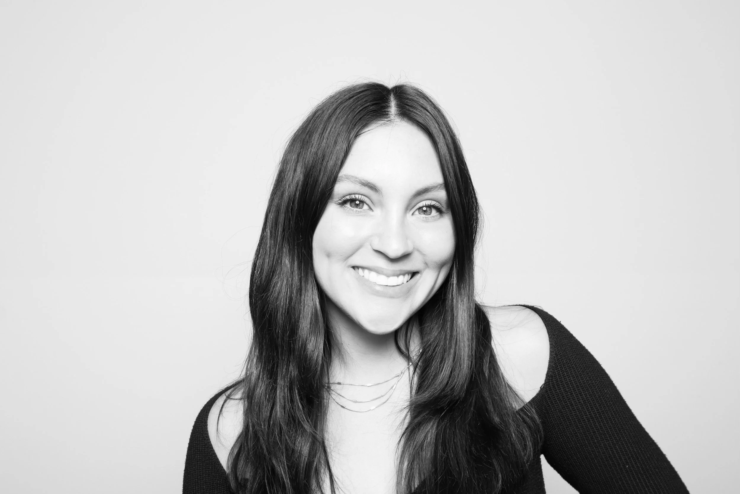 Black and white portrait of a smiling young woman with long dark hair, wearing a black top and layered necklaces, against a plain light background.