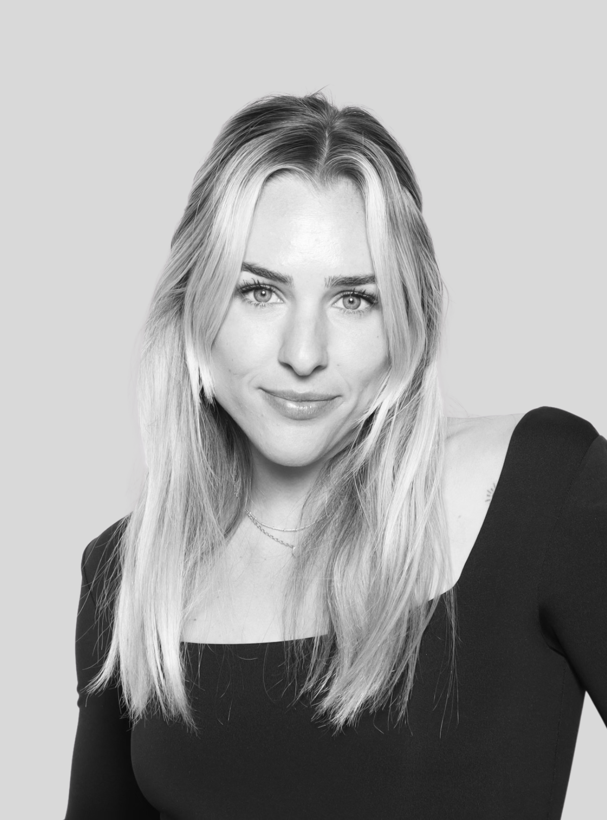 Black-and-white portrait of a young woman with long, wavy blonde hair, smiling slightly, wearing a black top with a wide neckline and layered necklaces, against a plain light background.