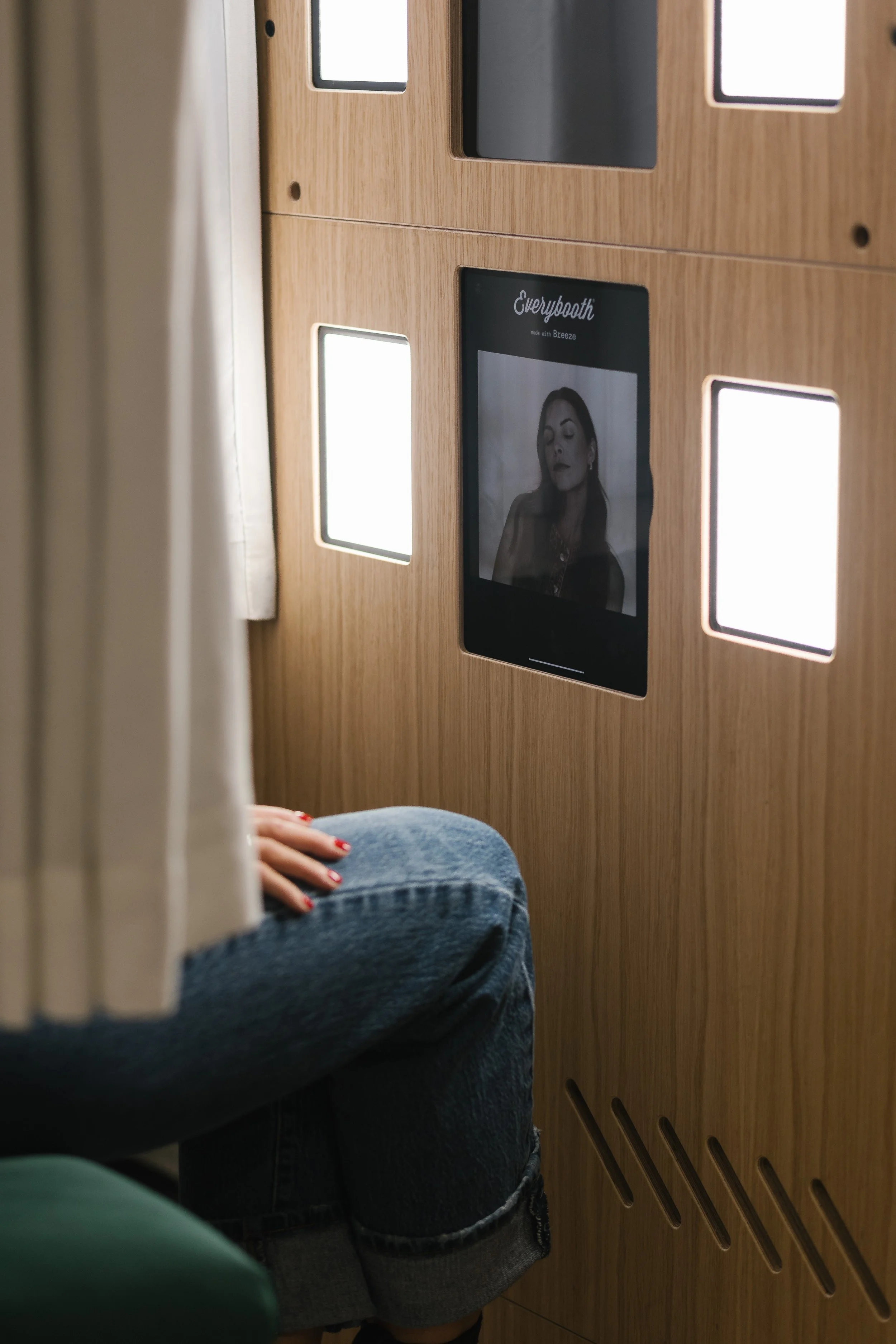 A person sitting in a wooden booth with a touchscreen monitor displaying a black-and-white photo of a woman with makeup, under the brand name 'Everybooth'.