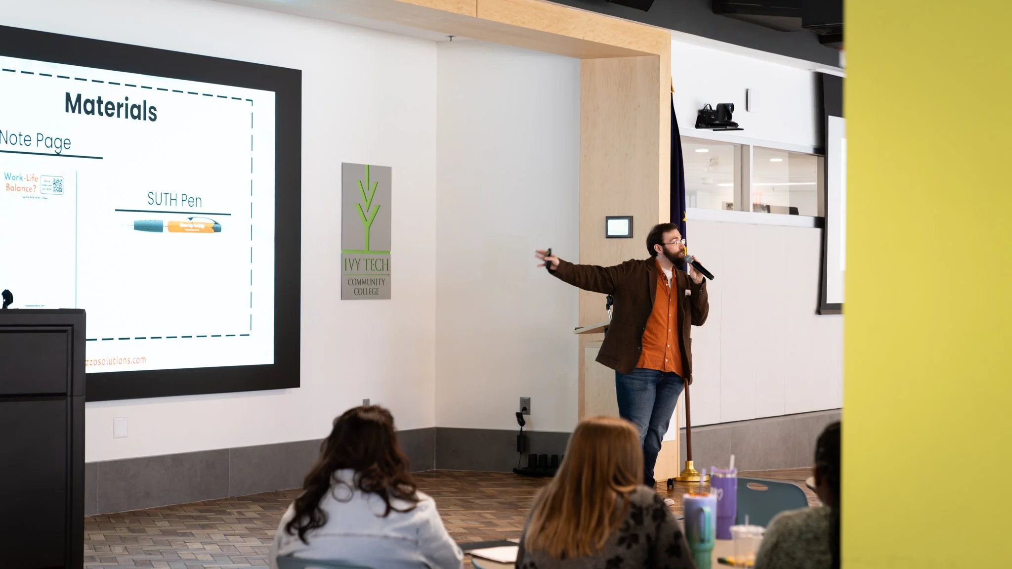 A man in glasses, a brown jacket, and jeans giving a presentation in a classroom or conference room with a microphone, a large screen displaying a slide with materials, and three women seated at a table in the foreground.