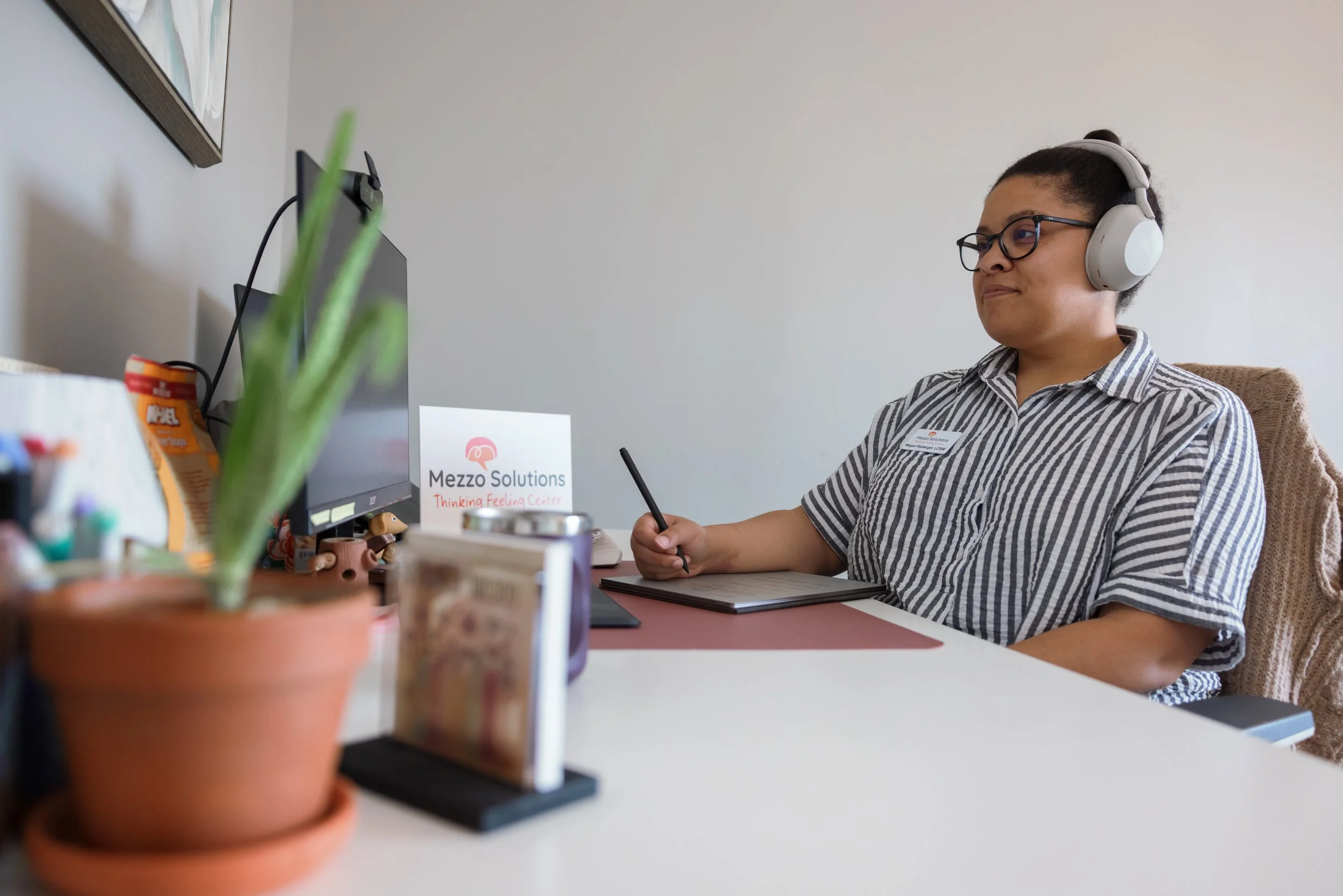 Woman wearing headphones and glasses, sitting at a desk with a tablet and pen, in front of a computer monitor.