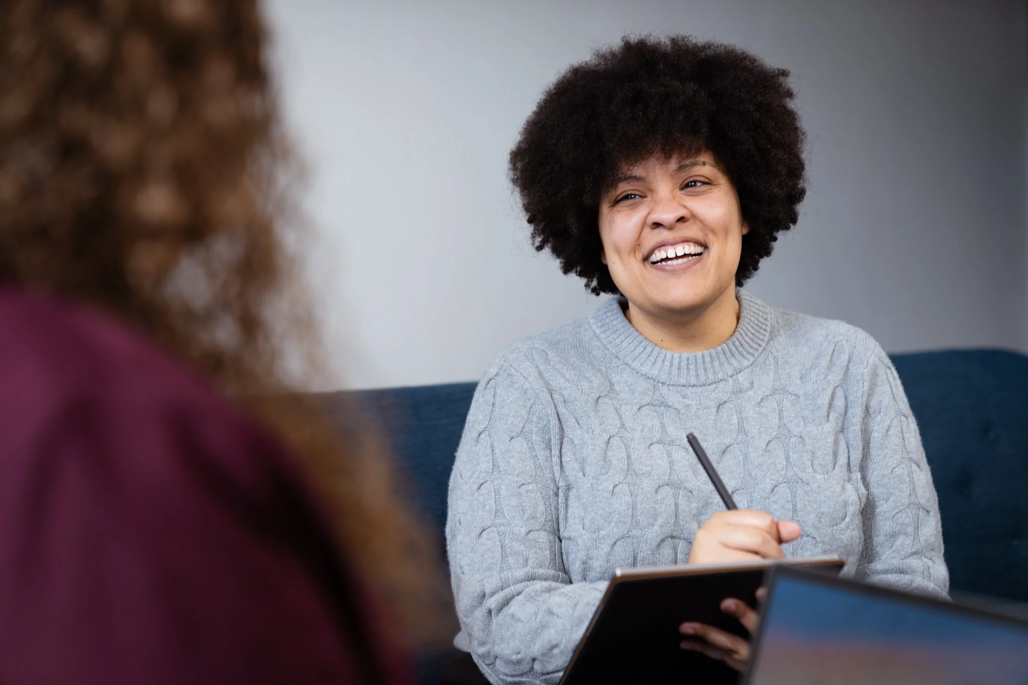 A woman with curly black hair, wearing a gray cable-knit sweater, smiling and holding a pen and notepad during a conversation.