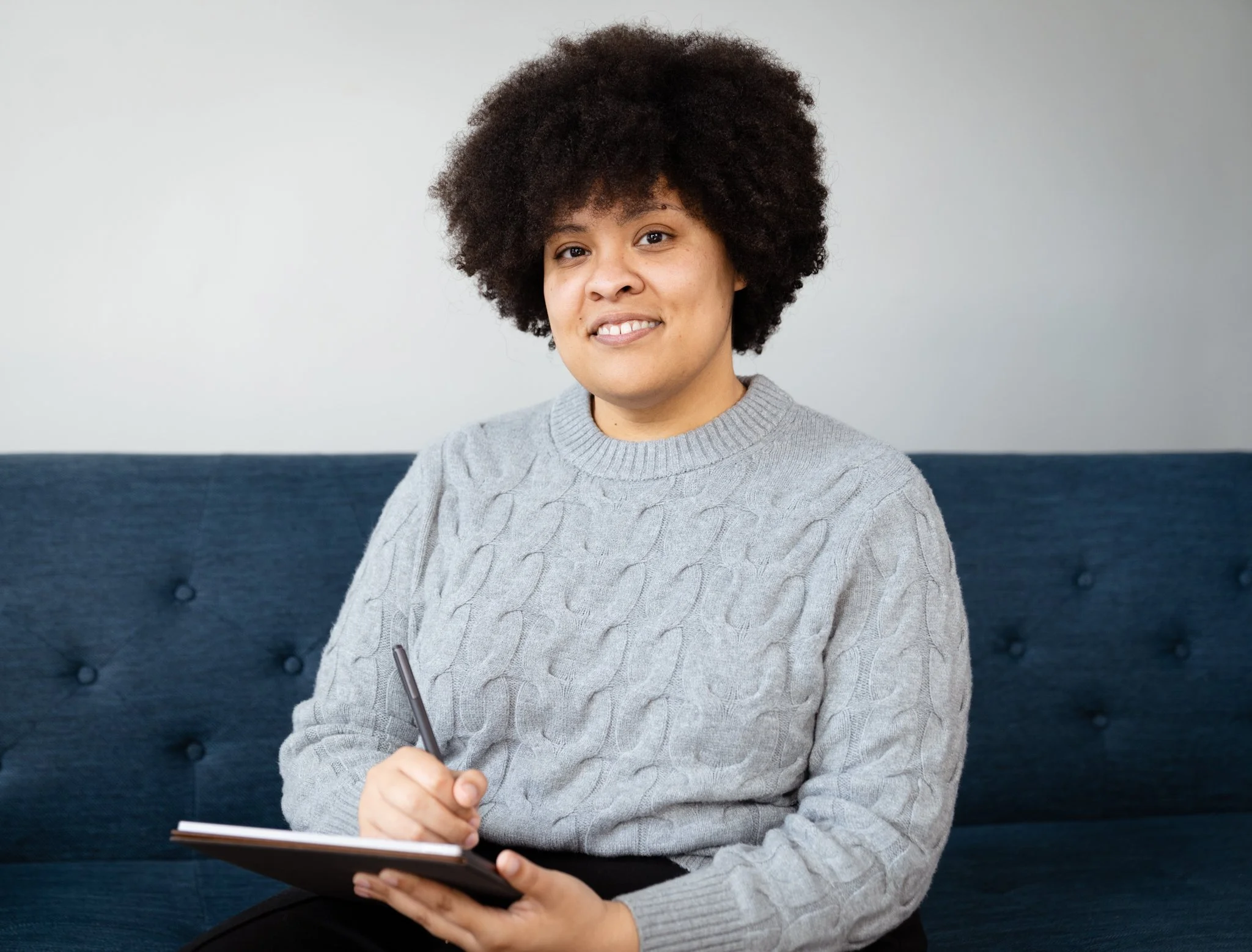 Woman with curly hair in a gray sweater sitting on a blue couch holding a notepad and pen.