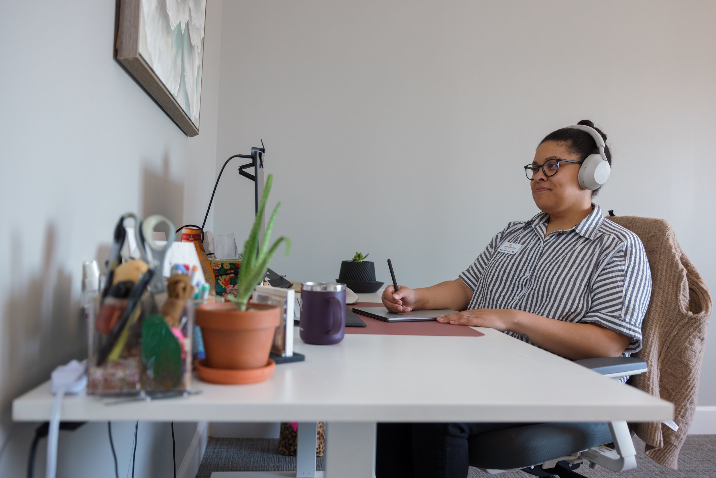 A woman with glasses and a striped shirt sitting at a desk, wearing headphones, working on a tablet or notebook in a home office with a white wall and decoration on the wall.