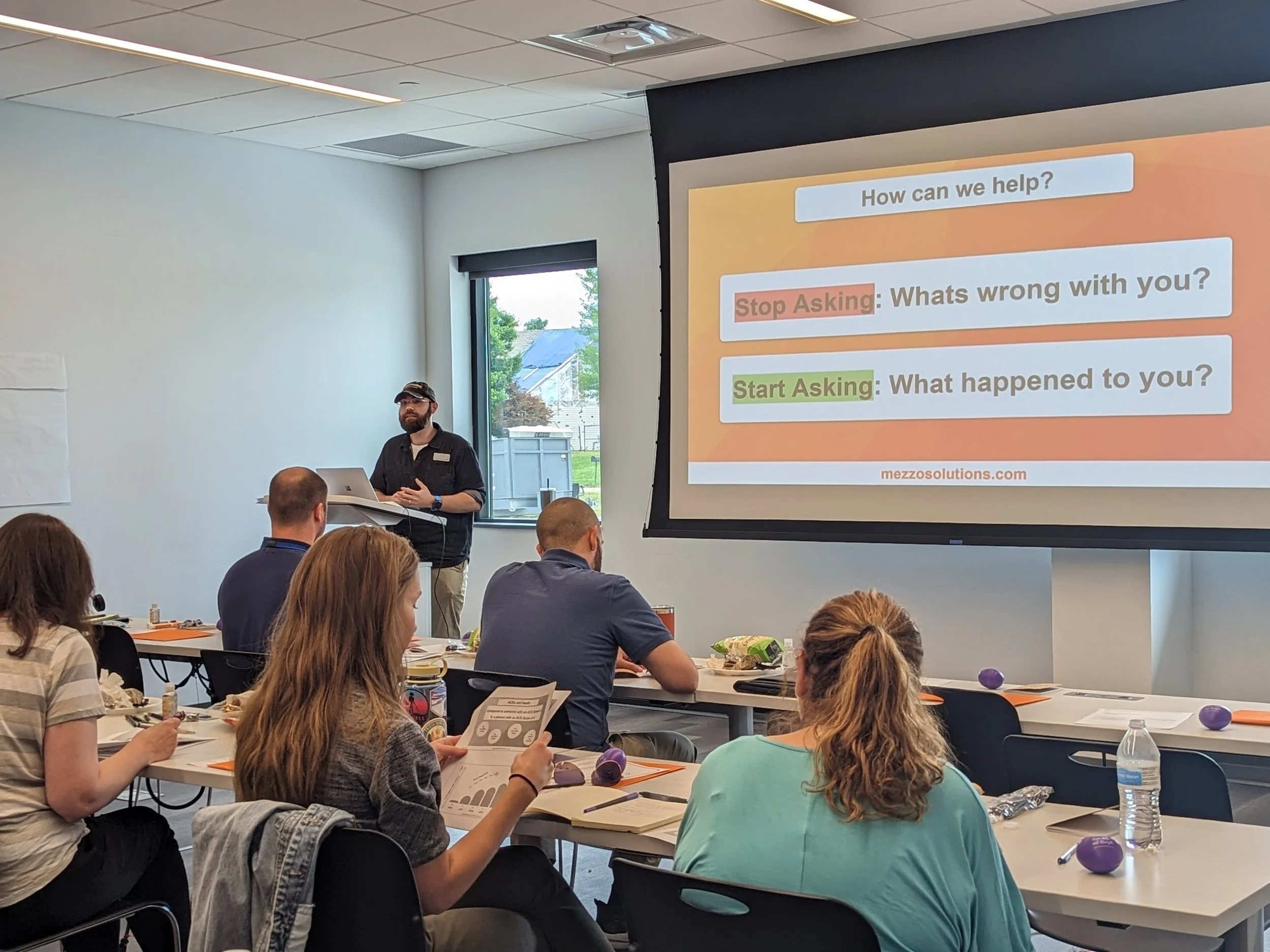 A man with a beard and glasses giving a presentation in front of a projector screen that displays motivational questions such as 'How can we help?', 'Stop Asking: What's wrong with you?', and 'Start Asking: What happened to you?'. The audience, mostly adults, sit at tables with papers, pens, water bottles, and purple stress balls in a bright conference room with windows.