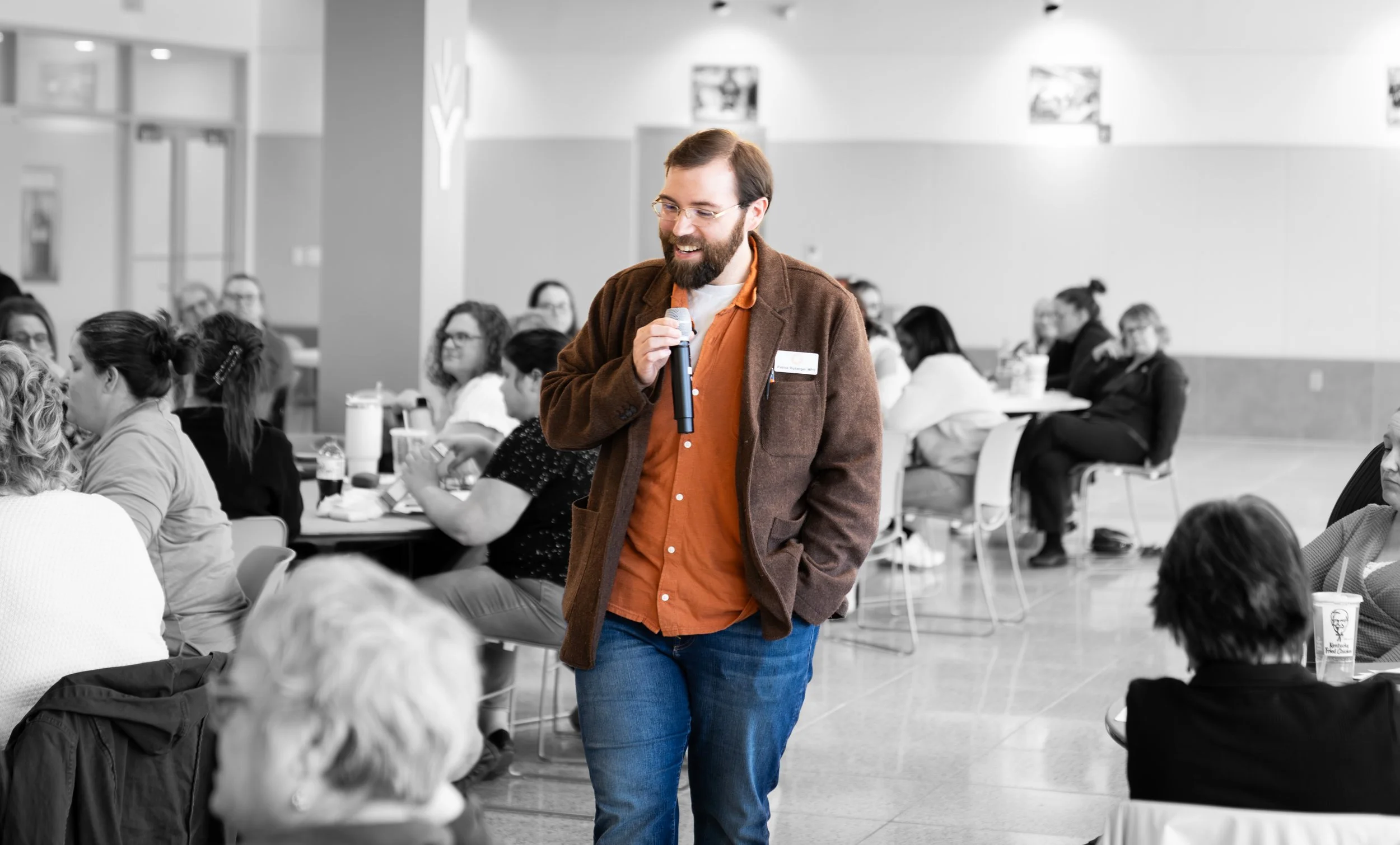 A man standing with a microphone in hand, speaking or presenting to a seated audience in a large, bright room. The image is in black and white, highlighting the man with a beard wearing glasses, a brown jacket, an orange shirt, and jeans, while the audience appears to be listening attentively.