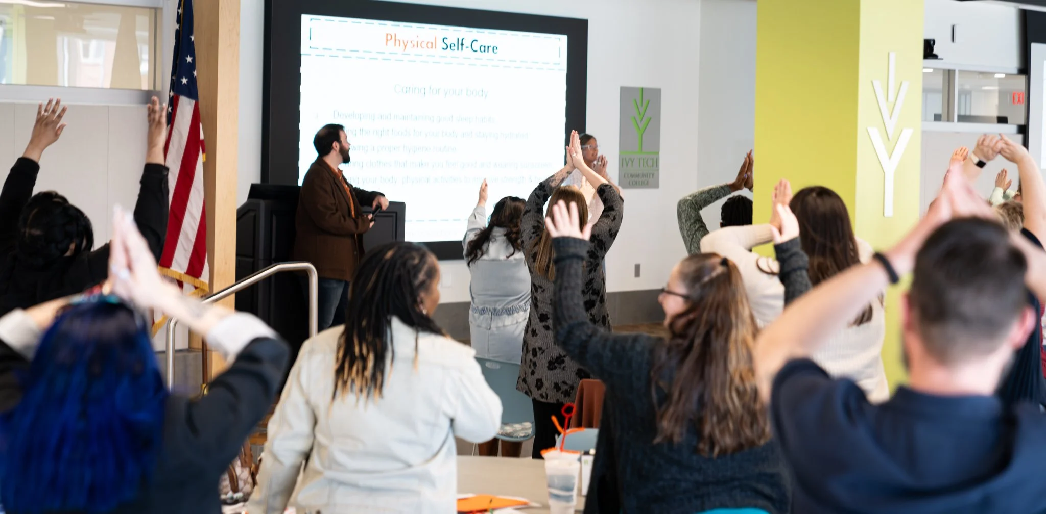 People participating in a workshop or seminar, raising their hands in response to a presenter at the front who is standing near a large screen displaying a presentation about physical self-care.