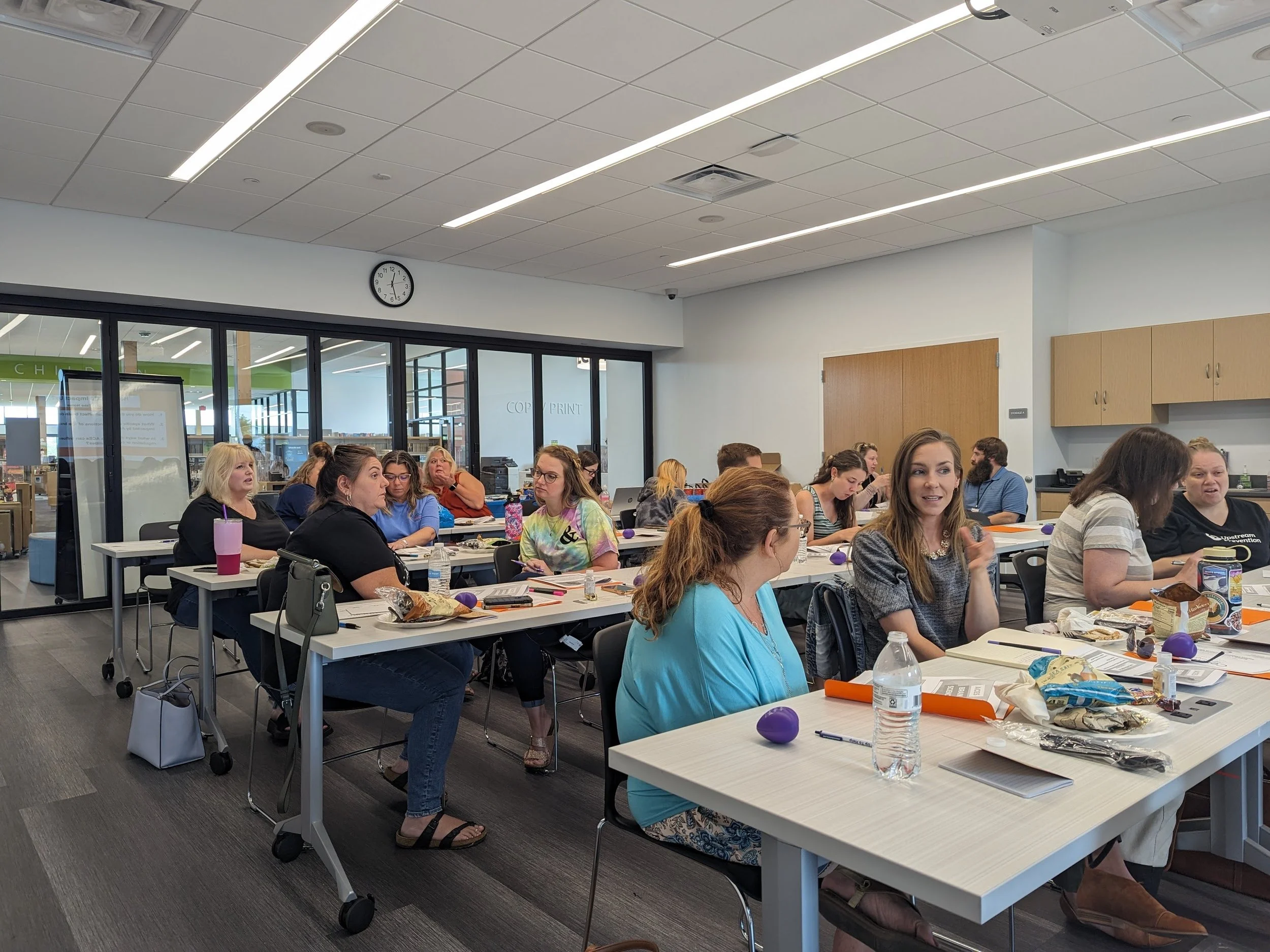 A group of diverse women sitting at long tables in a conference room, engaged in discussion. The room has a white ceiling with linear lights, a wall clock showing 2:11, and large glass windows and doors leading outside. Food and drinks are on the tables.