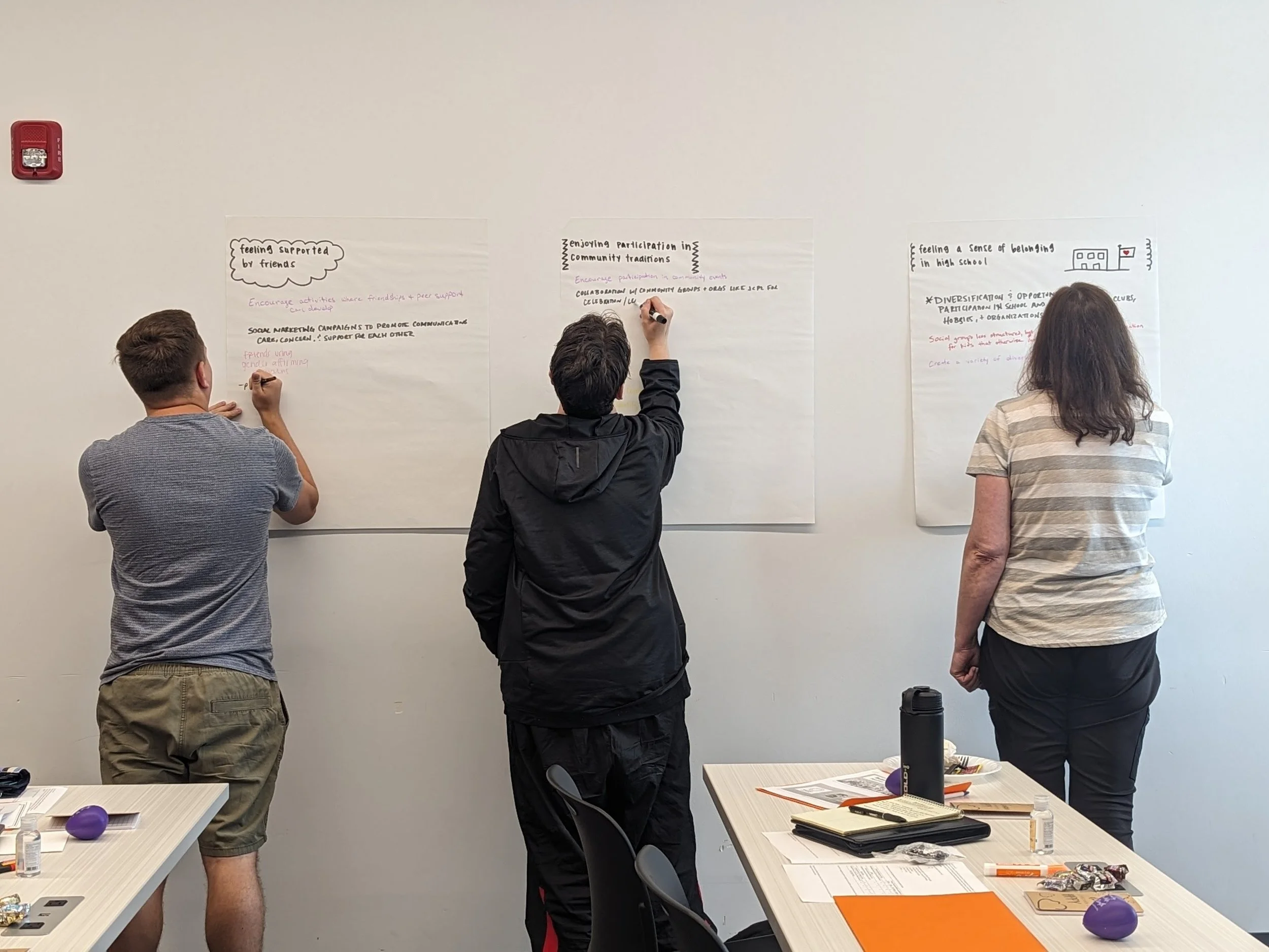 Three people standing at a whiteboard with handwritten notes and diagrams in a classroom or meeting room. Two people are writing on the whiteboard, while a woman looks on. There are tables in the foreground with papers, notebooks, pens, snacks, and drinks.