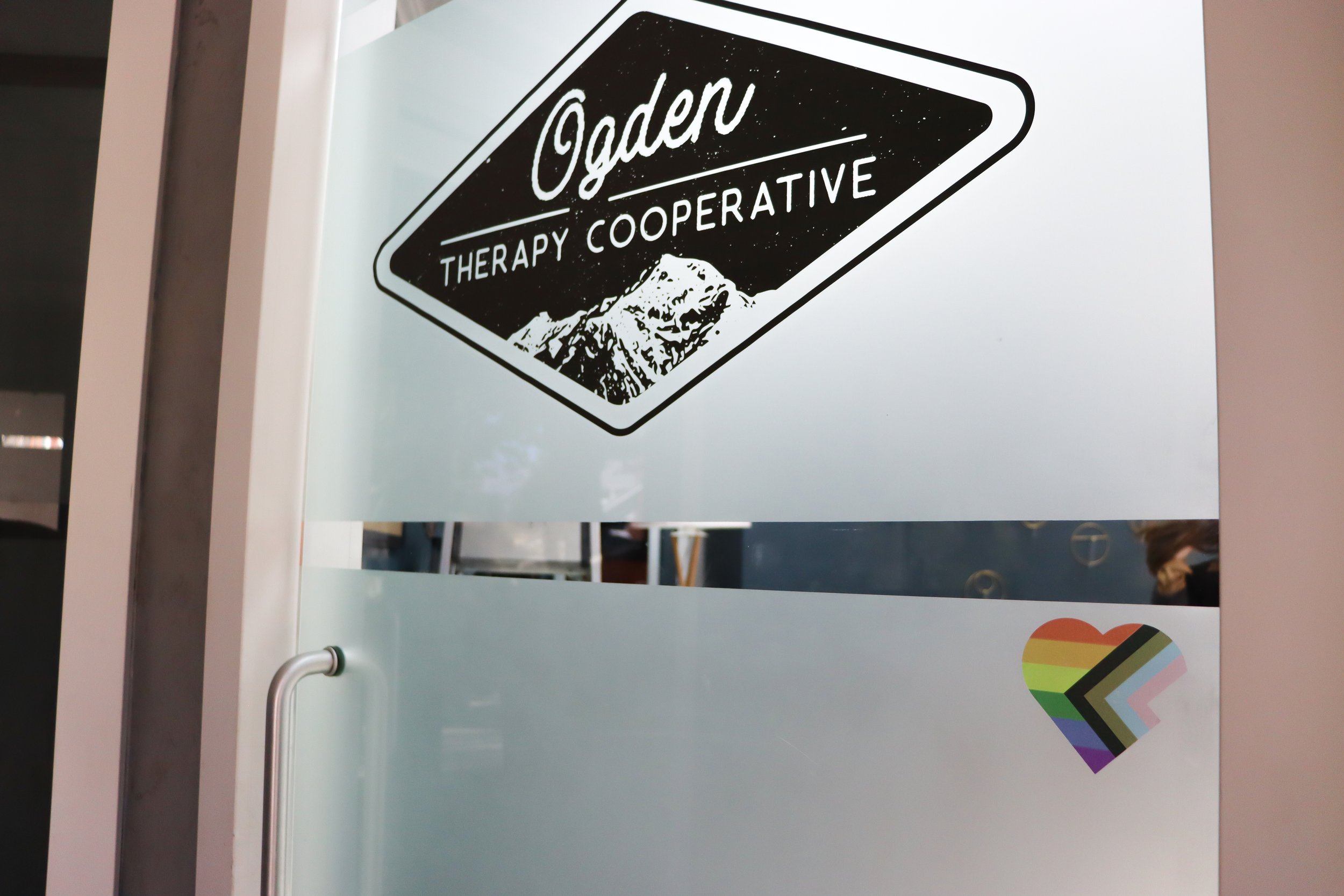 Glass frosted door with lgbtqia+ heart on corner and logo for Ogden therapy cooperative in black on the door