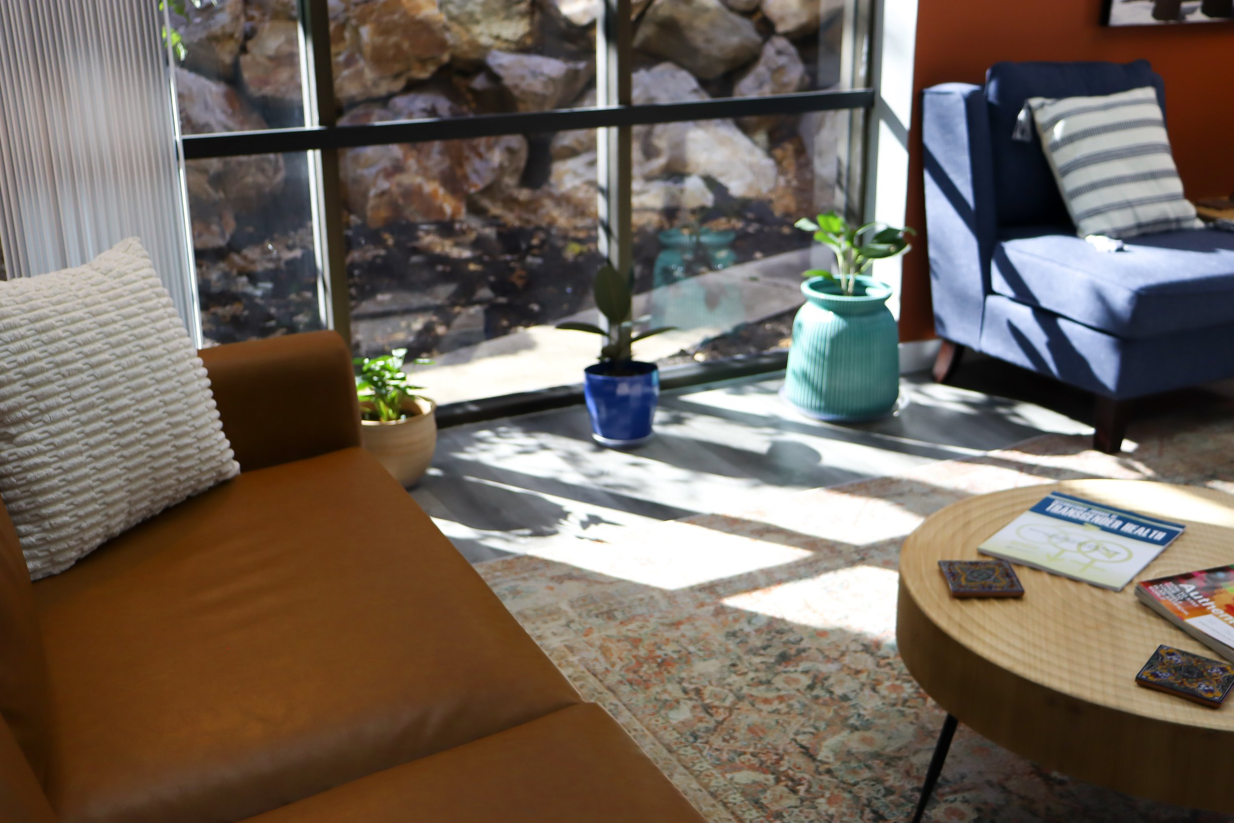 Office rental space lobby with large window and sun streaming in on plants. Orange sofa to left, coffee table with magazines, and lounge chairs.