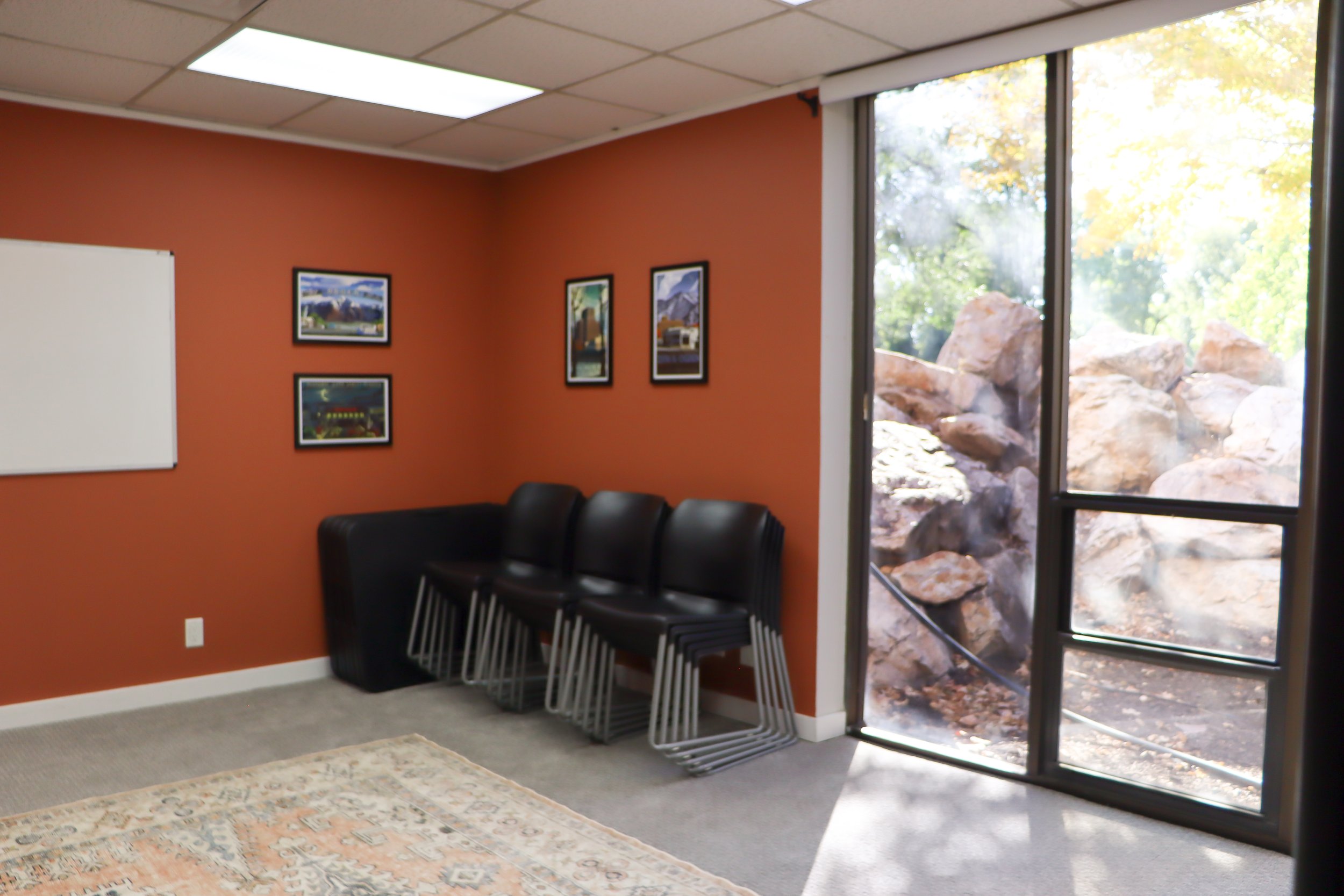 Group therapy room with chairs stacked in corner, large window, whiteboard, and burnt orange walls