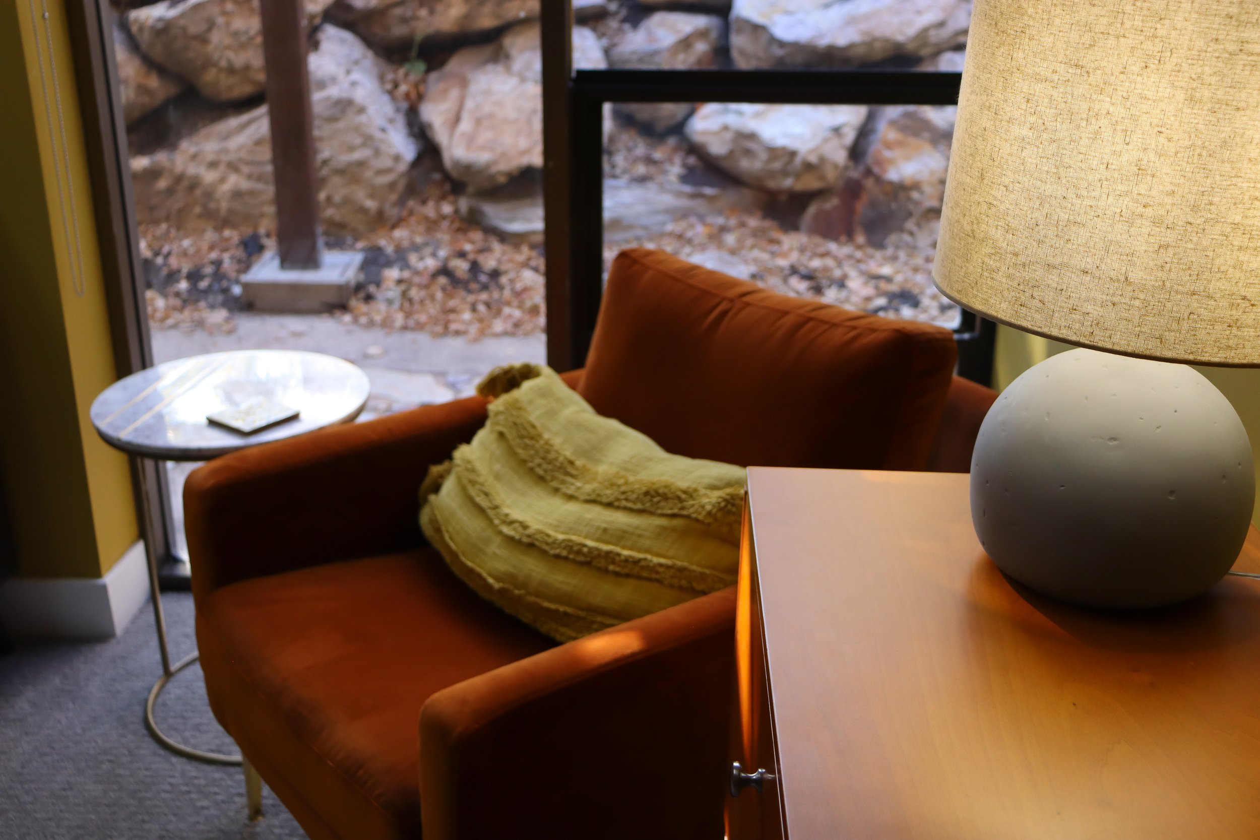 Modern orange velvet chair in therapy office with large window behind it and a warm lamp lighting the space