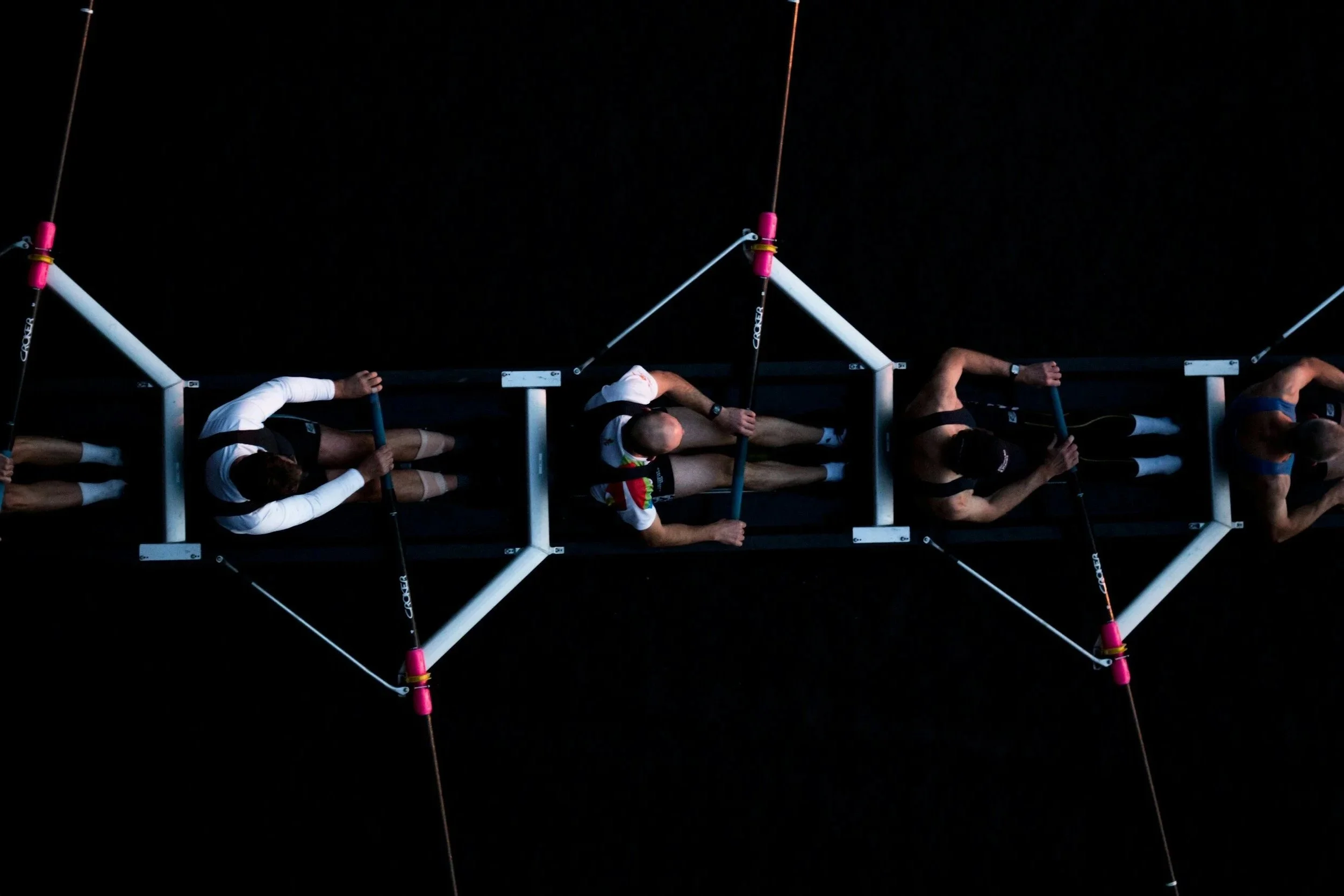 Four rowers in a boat during a race, viewed from above against dark water.
