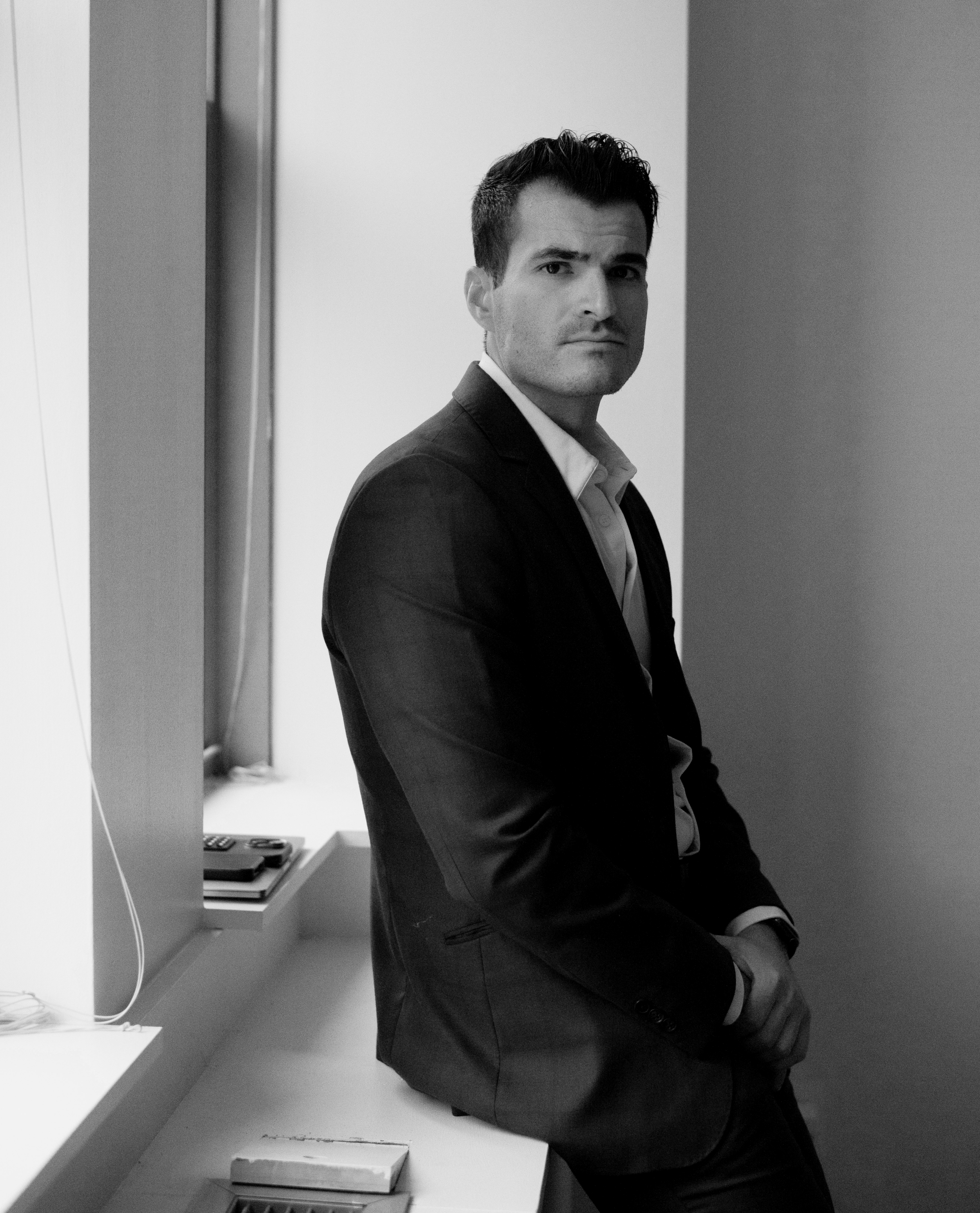 A man in a suit leaning against a wall in an office, looking serious.