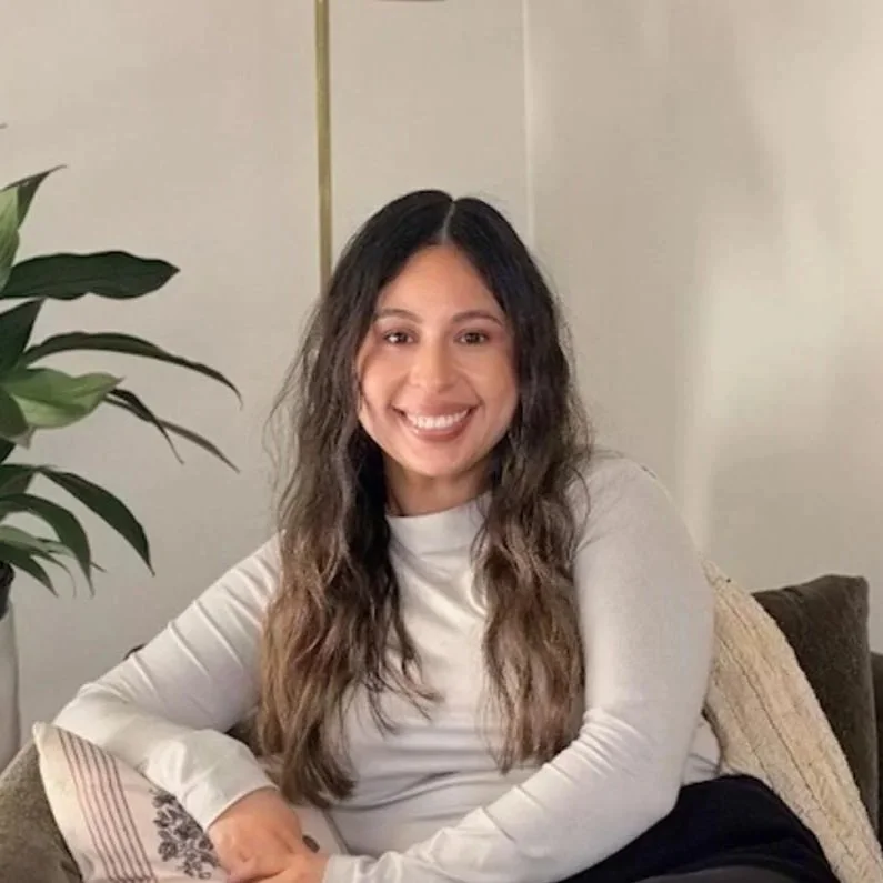 A woman with long, wavy dark hair smiling and sitting on a couch in a living room.