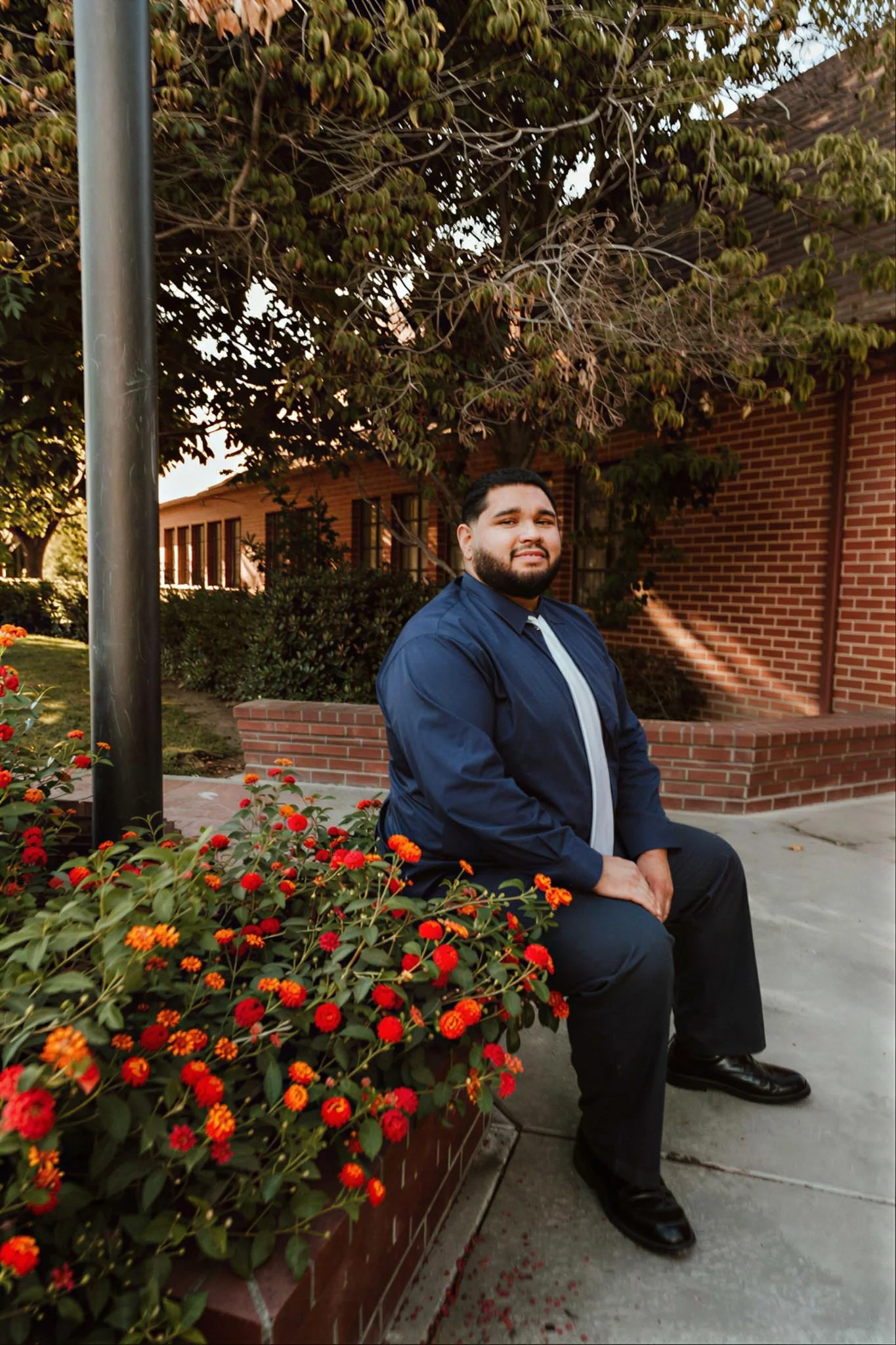 A man in a dark blue shirt, gray tie, and black pants sitting on a bench next to a flowering bush with red and orange flowers, outdoors near a brick building and green trees.