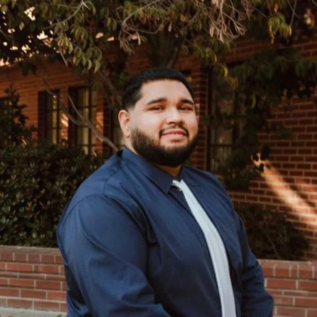 A man with dark hair and a beard wearing a blue jacket stands outdoors in front of a brick building with trees in the background.