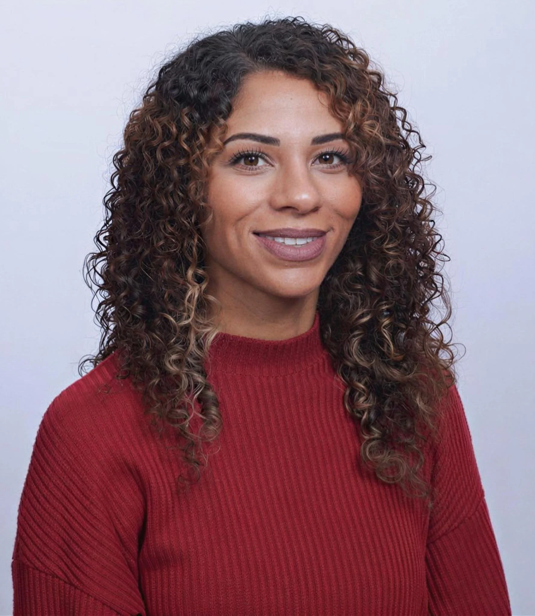 A woman with curly hair and light brown skin smiling, wearing a red sweater against a plain light background.