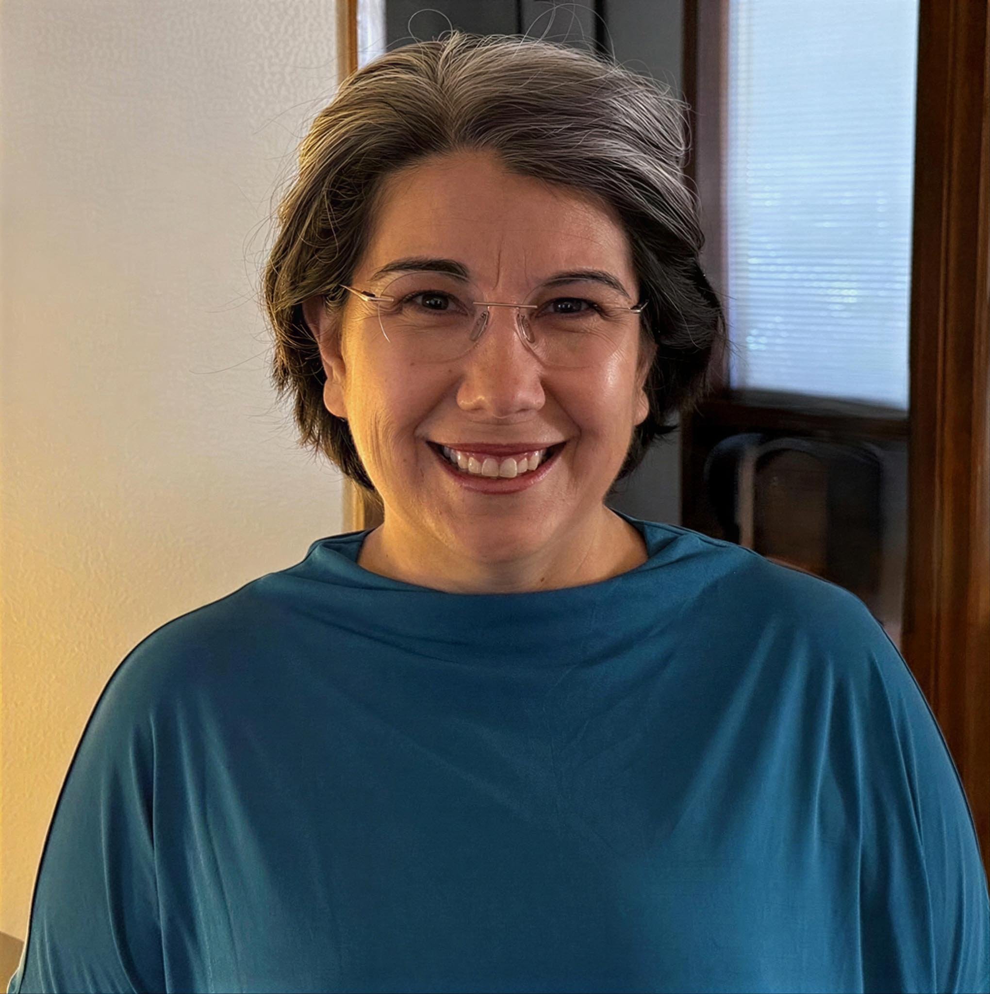 A woman with short gray hair, wearing glasses and a blue top, smiling indoors with a wooden door and window in the background.