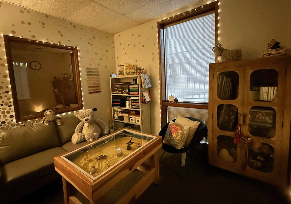 A cozy living room with a sofa, several teddy bears, a glass-topped display table, a black chair with a colorful pillow, a wooden cabinet with glass doors, a bookshelf filled with books and decorative items, a wall mirror with string lights, a window with blinds, and warm ambient lighting.