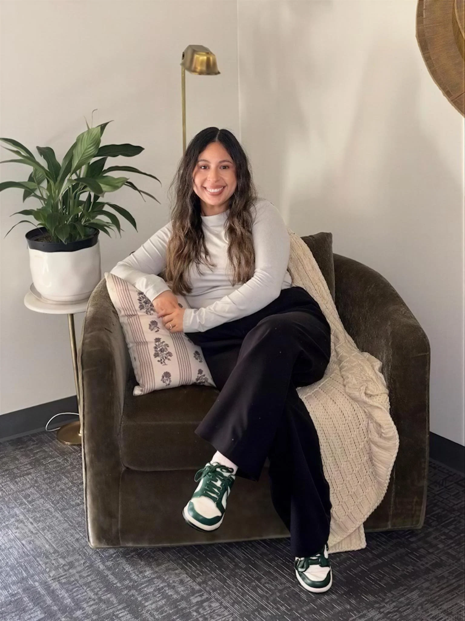 A young woman with long, wavy hair, sitting on a brown velvet armchair, smiling. She's wearing a white long-sleeve top, black pants, and green-and-white sneakers. Next to her is a side table with a potted green plant, and a gold-colored floor lamp.