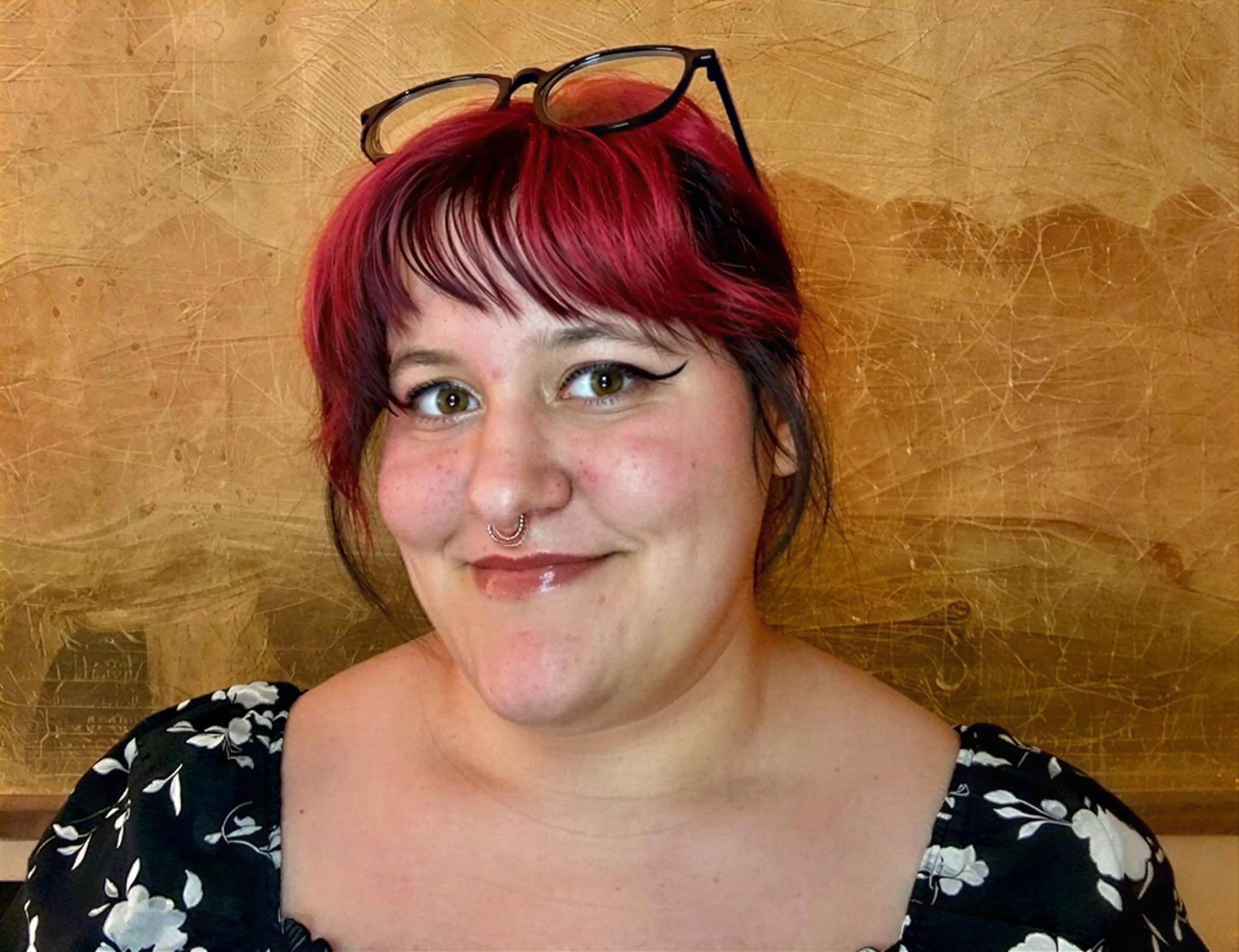 A woman with short red and black hair, glasses on her head, and a septum piercing smiling in front of a wooden background.