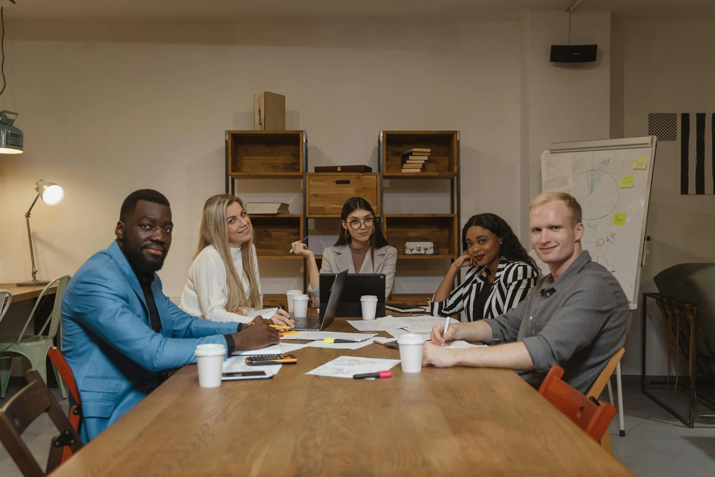Five diverse young adults socializing in a modern room with green plants, two women sitting on chairs and three people standing, engaged in conversation.