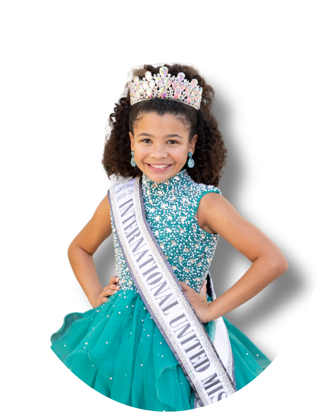 Young girl with a crown, earrings, and a sash that reads "International United Miss" wearing a glittering turquoise gown with a tulle skirt and pose with her hands on her hips, smiling.