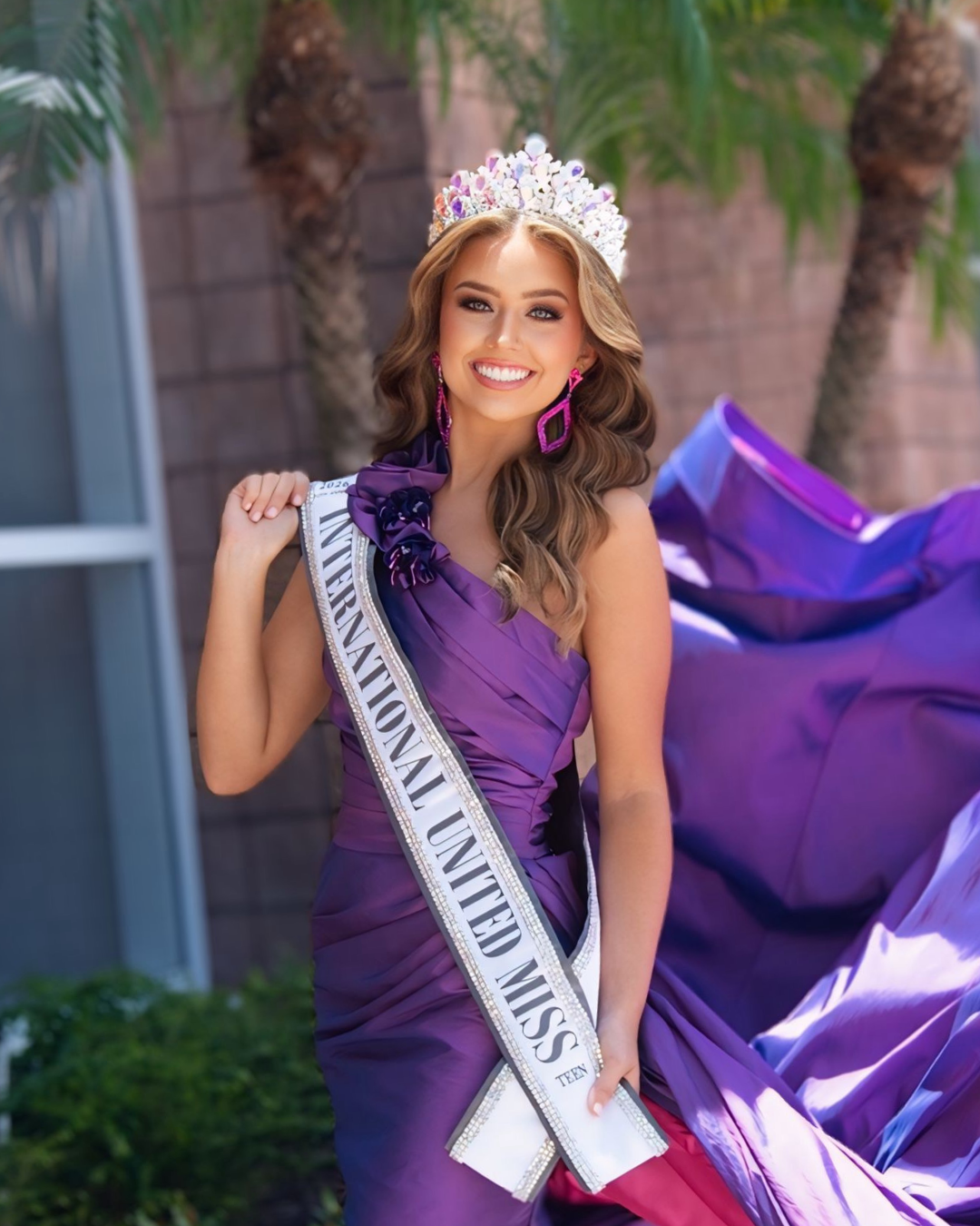 A young woman wearing a tiara, sash, and a pink gown with intricate beadwork, smiling confidently with one hand on her hip, against a black background.